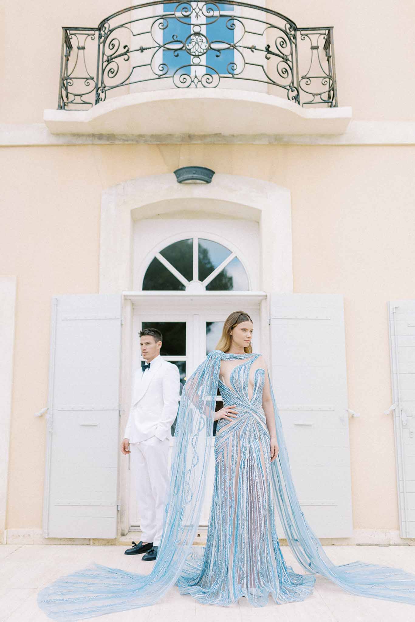 Bride in beaded blue gown with floor-length cape groom in white tuxedo before peach-toned chateau with wrought-iron balcony