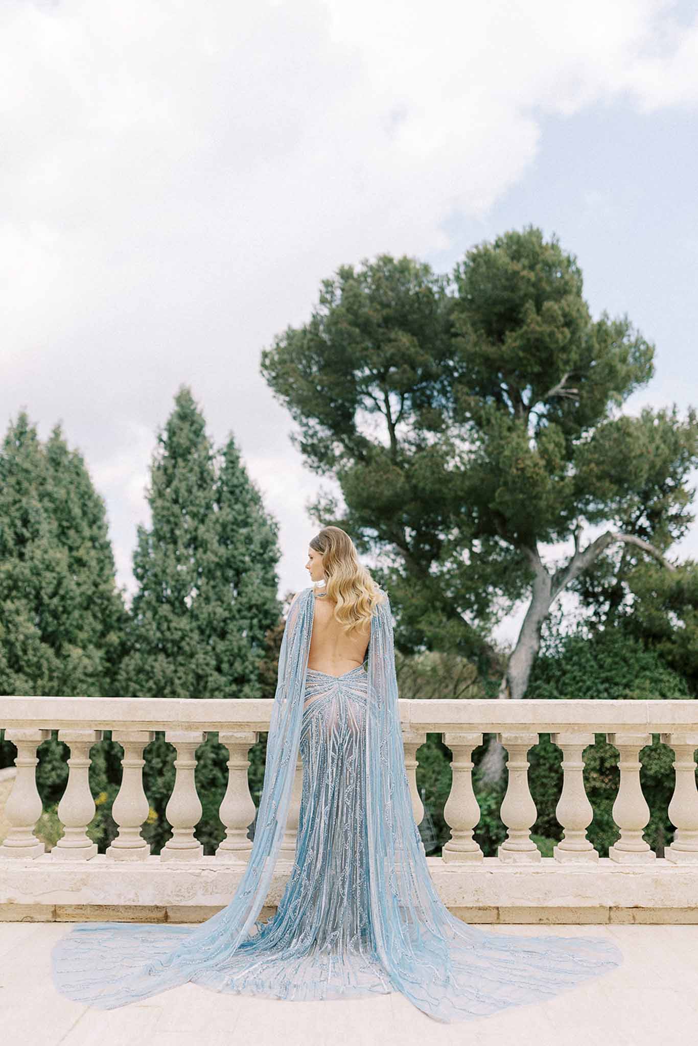 Bride photographed from behind wearing pale blue embellished gown with sheer cape on stone balustrade terrace