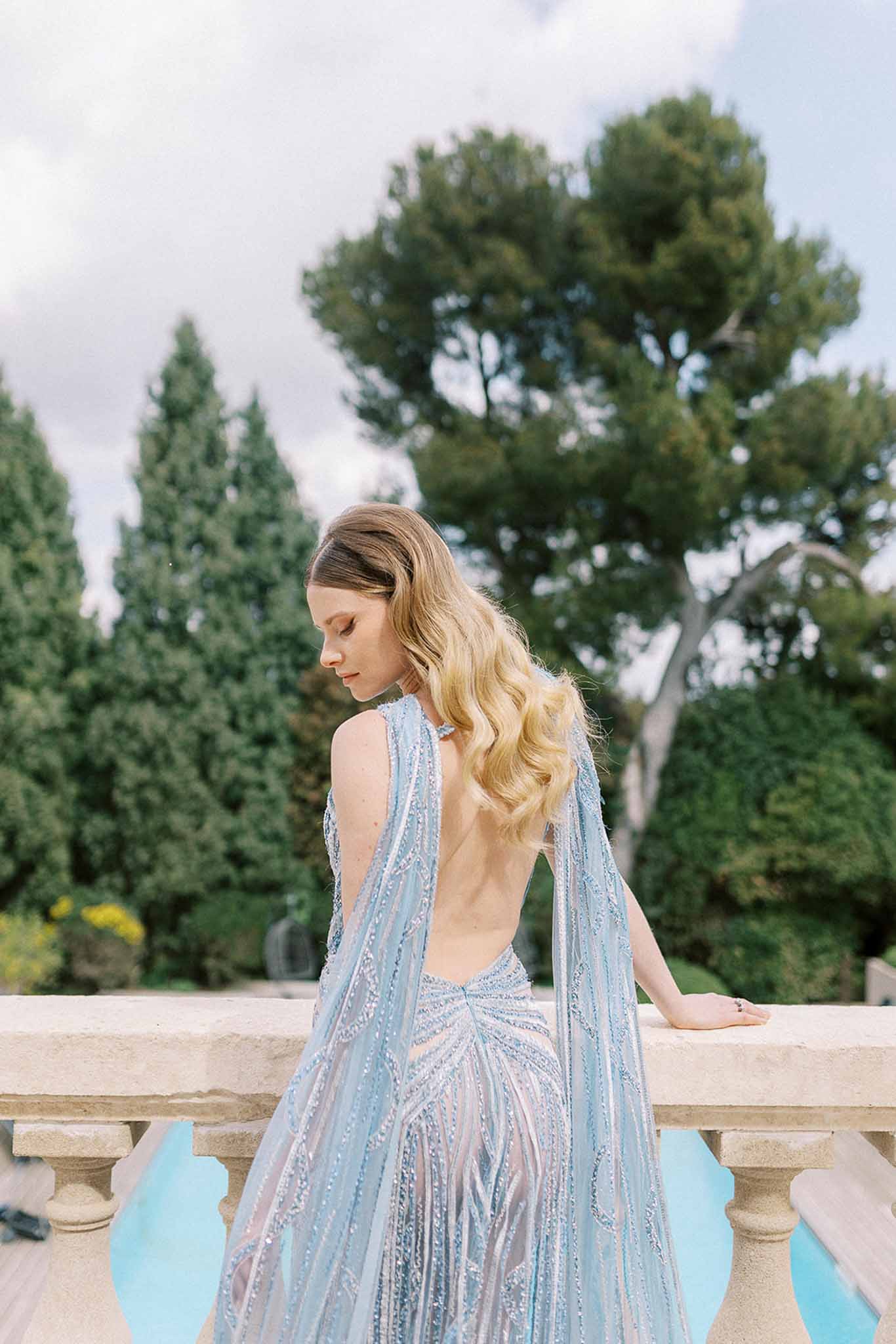 Bride from behind in pale blue beaded open-back gown with cape detail leaning on stone balustrade above pool