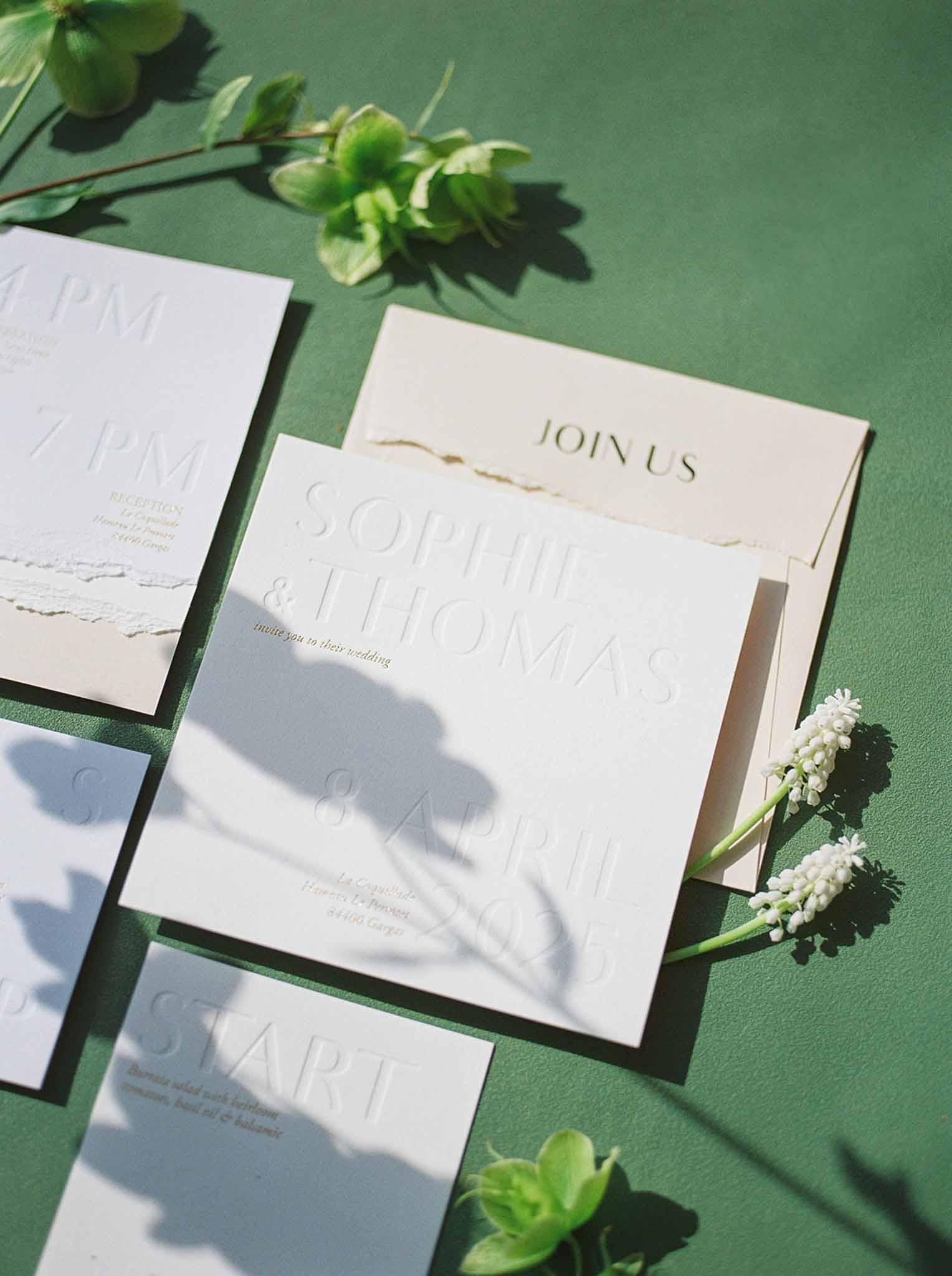 Wedding stationery flat lay with white embossed invitation suite on green surface with white muscari stems