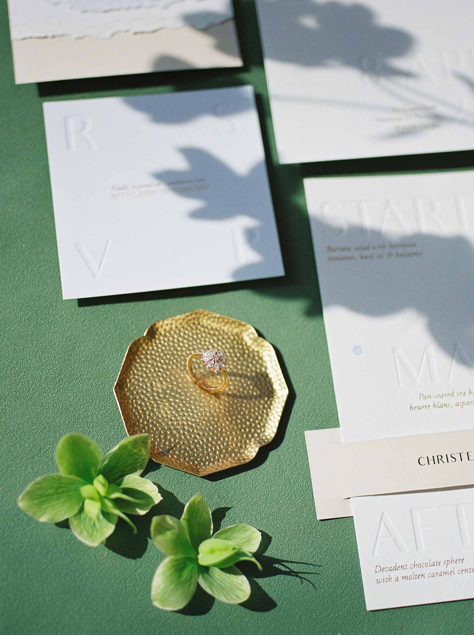 Flat lay of white monogrammed wedding stationery suite with gold engagement ring and green hellebore stems on dark green s...