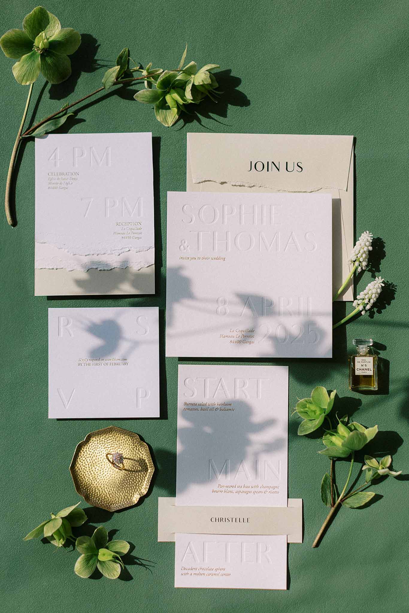 Wedding stationery flat lay with embossed white suite, gold ring dish, Chanel perfume on green background