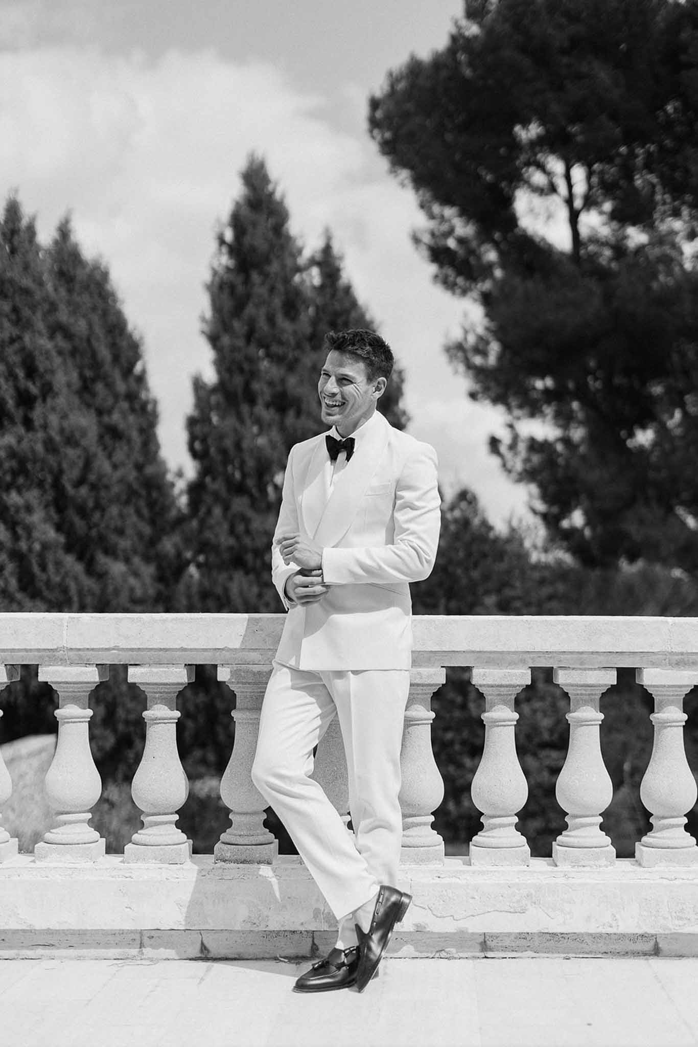 Black and white groom in white tuxedo laughing and adjusting cufflinks on chateau terrace balustrade