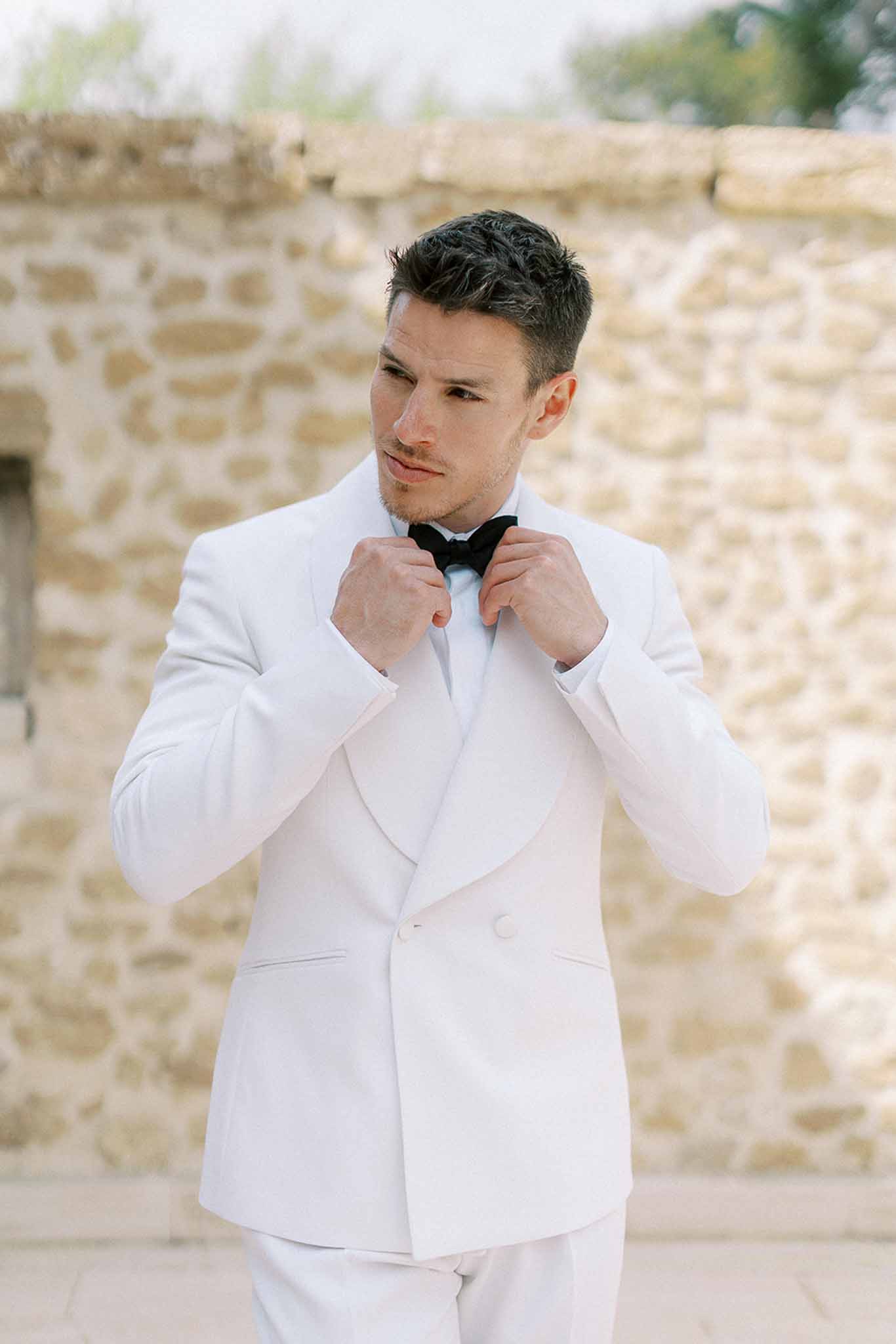 Groom adjusting black bow tie wearing white double-breasted tuxedo jacket against limestone wall