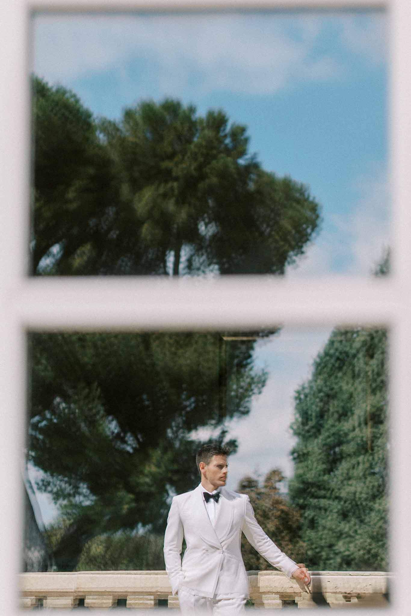 Groom in white suit and black bow tie photographed through a window frame on a chateau terrace