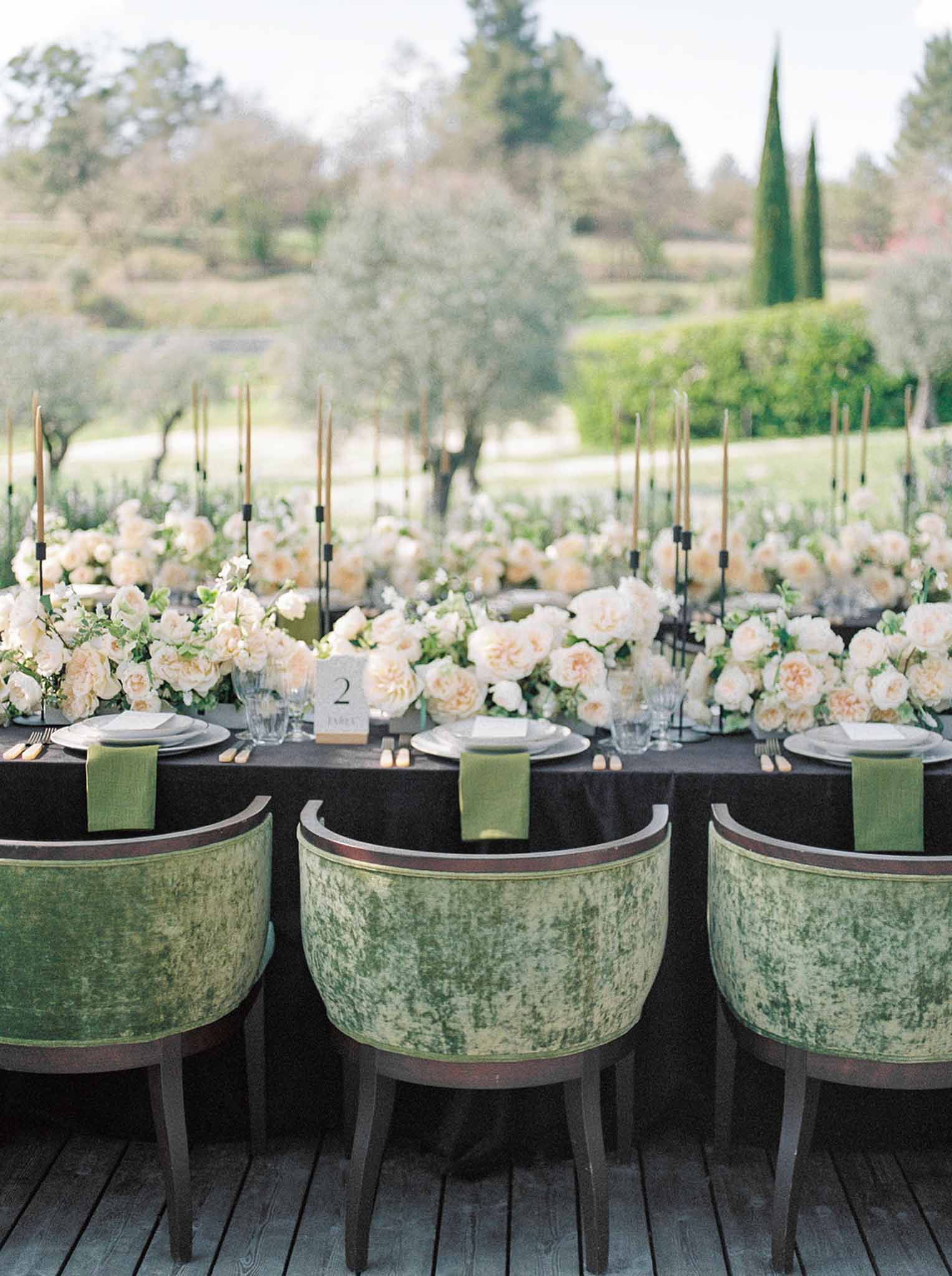 Reception tablescape with cream and blush rose runner, gold taper candles, and green velvet barrel-back chairs on wooden deck