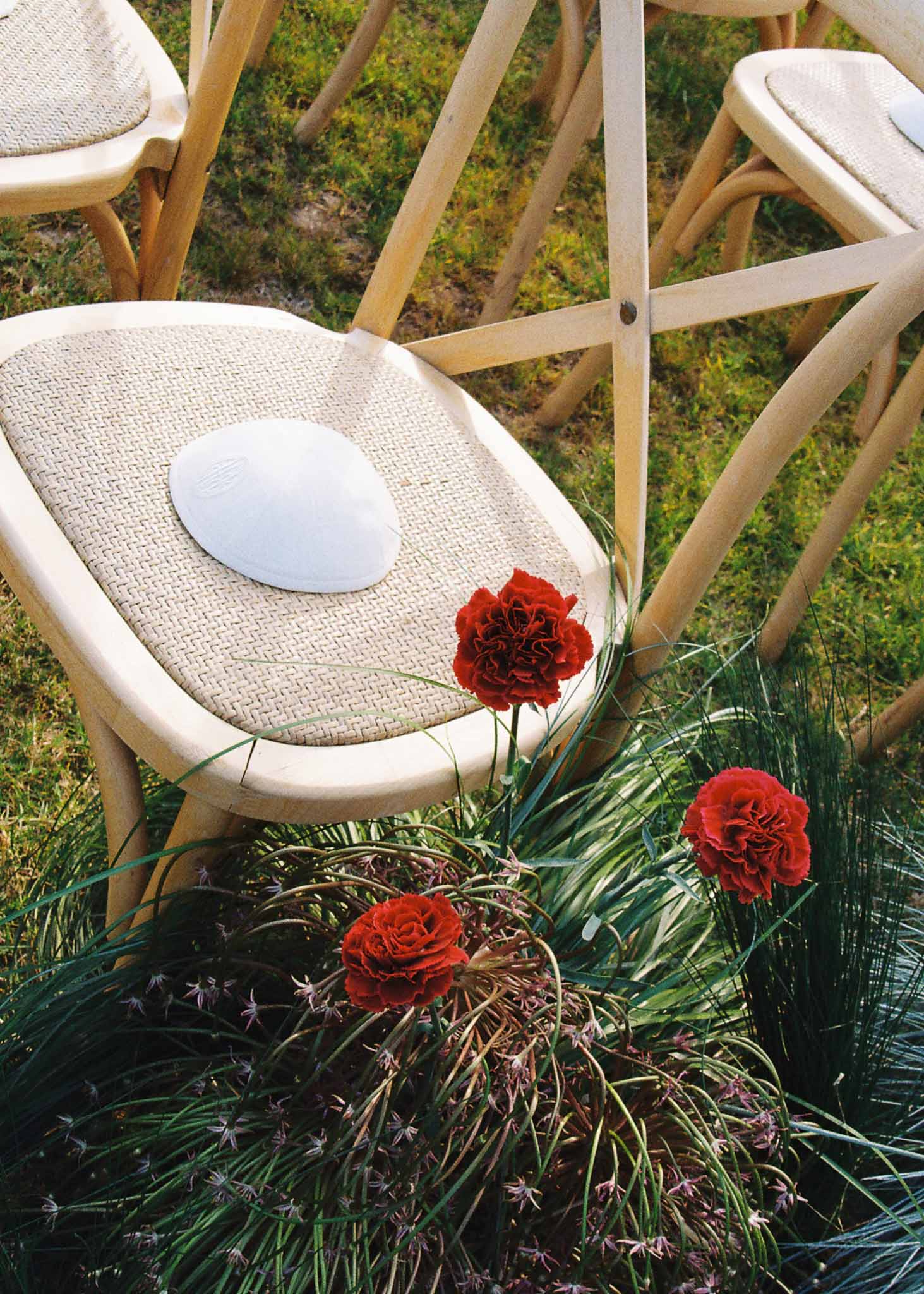 Red carnation and ornamental grass aisle arrangement beside crossback ceremony chair on lawn