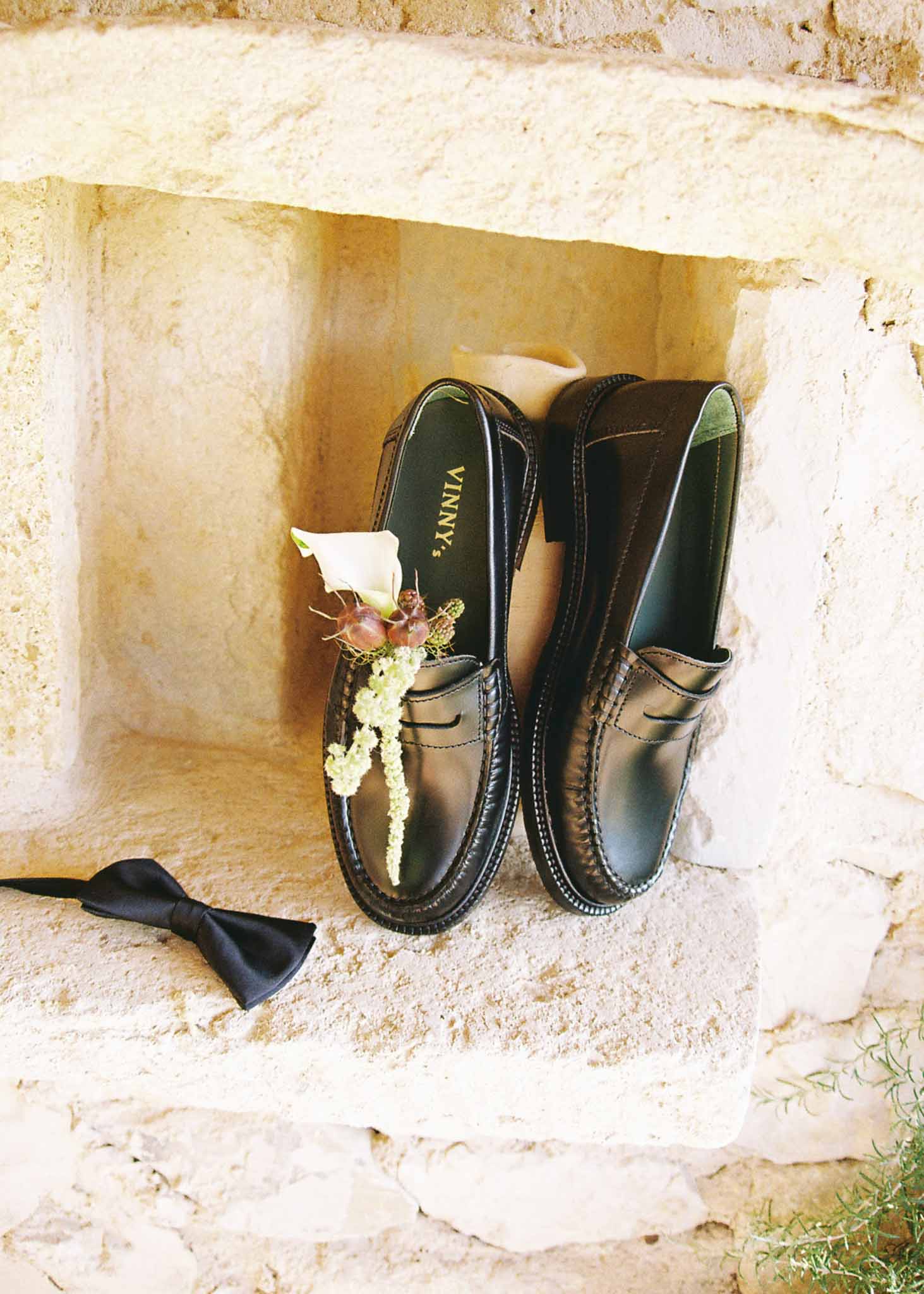 Groom accessories in limestone wall niche with black leather loafers, silk bow tie, and white calla lily boutonniere