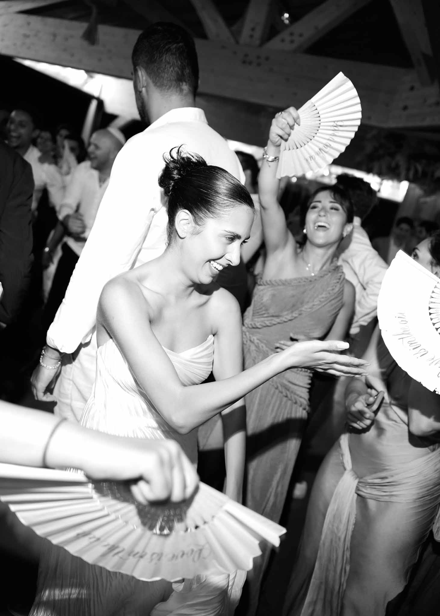 Black and white candid of guests dancing with personalized paper fans under exposed beam ceiling
