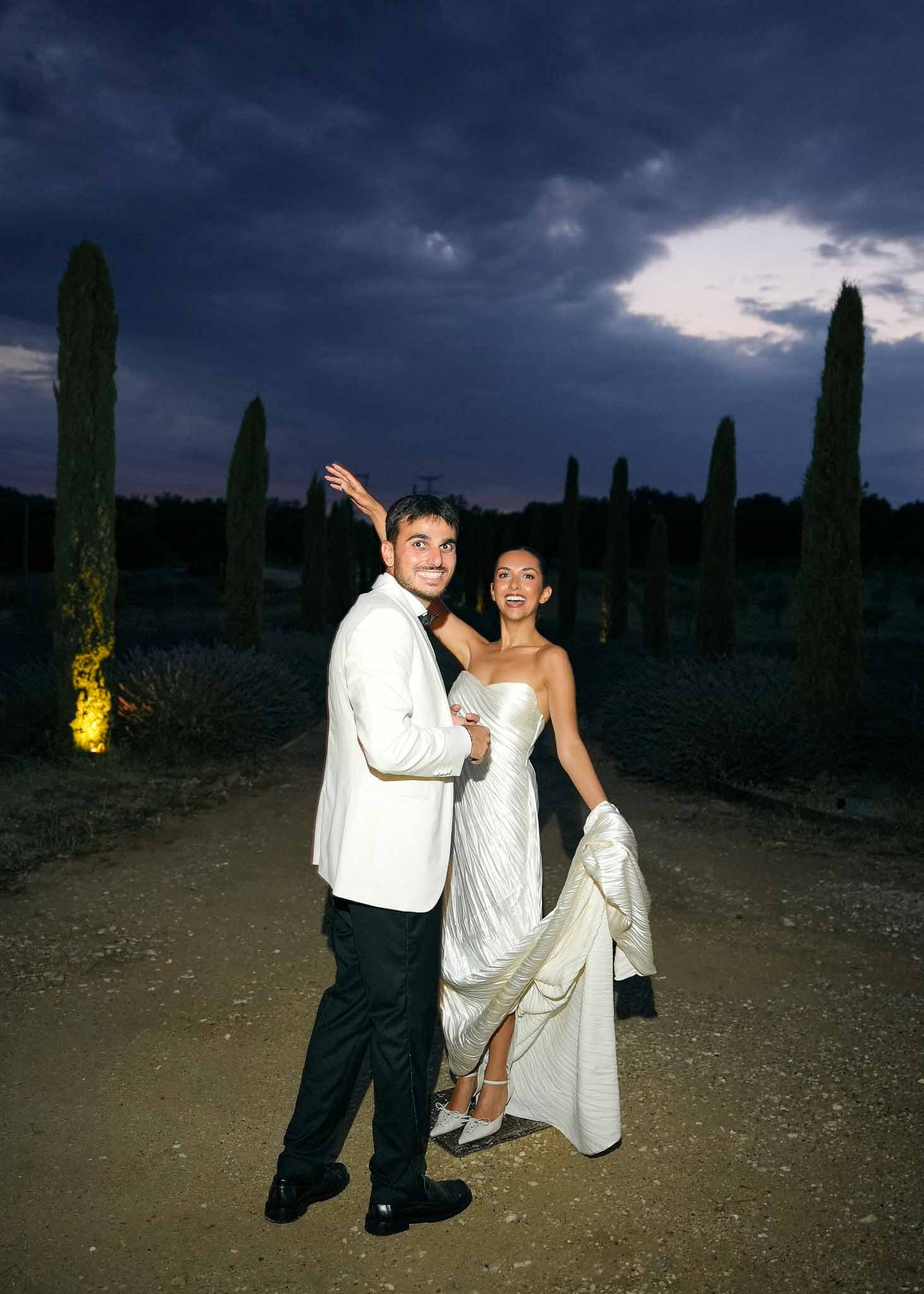 Playful couple on cypress-lined gravel path at dusk with amber uplighting, bride in silver-white draped gown
