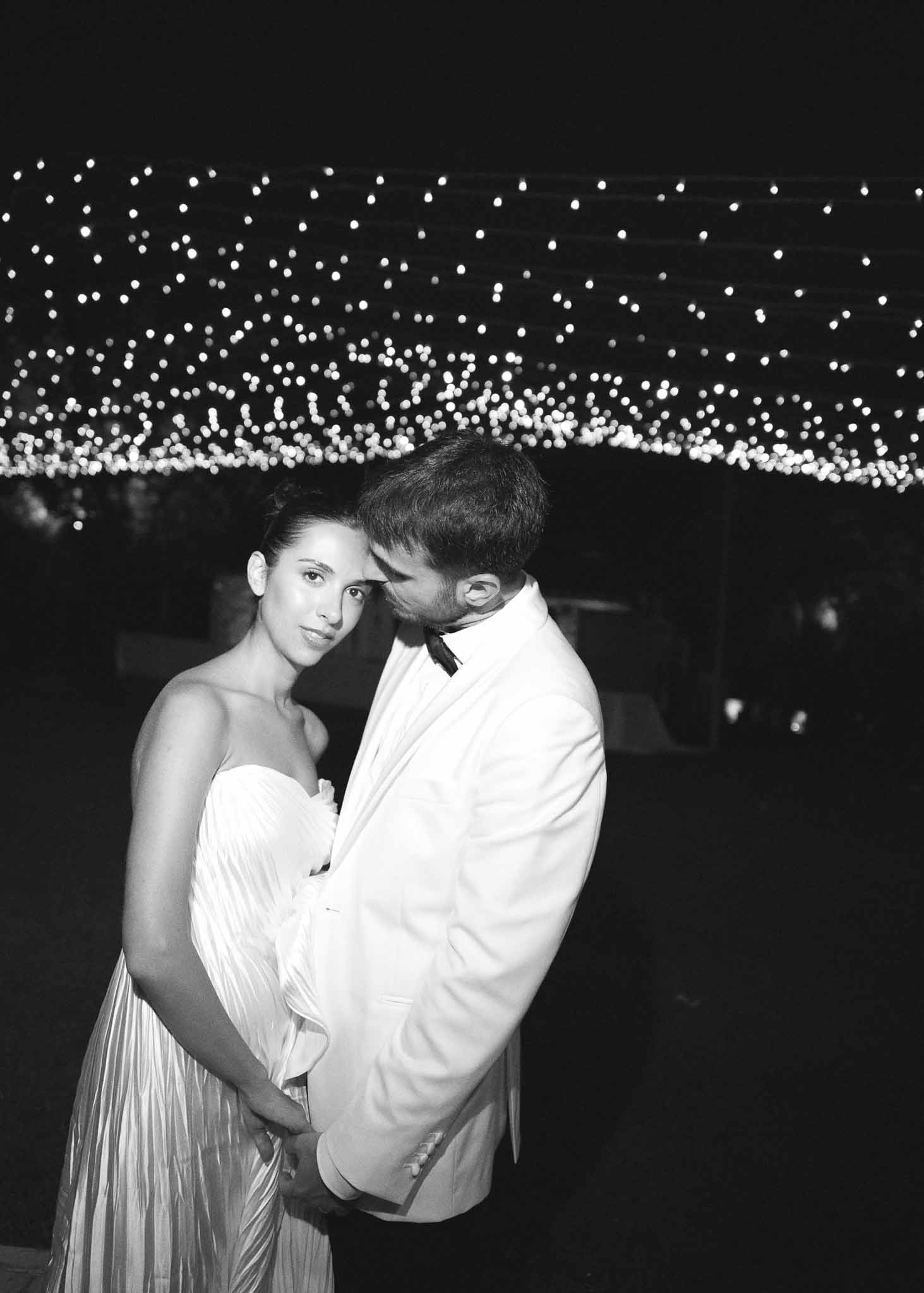 Black and white nighttime portrait of couple embracing under fairy light bokeh canopy
