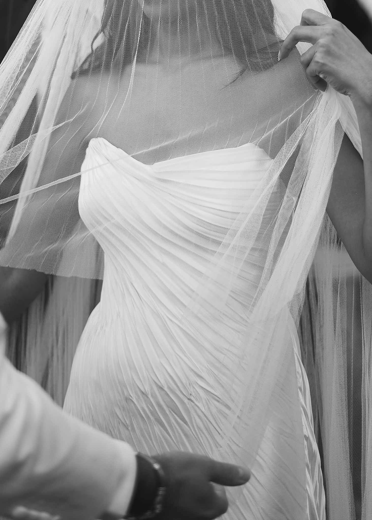 Black and white close-up of pleated strapless bodice and tulle veil being arranged by multiple hands
