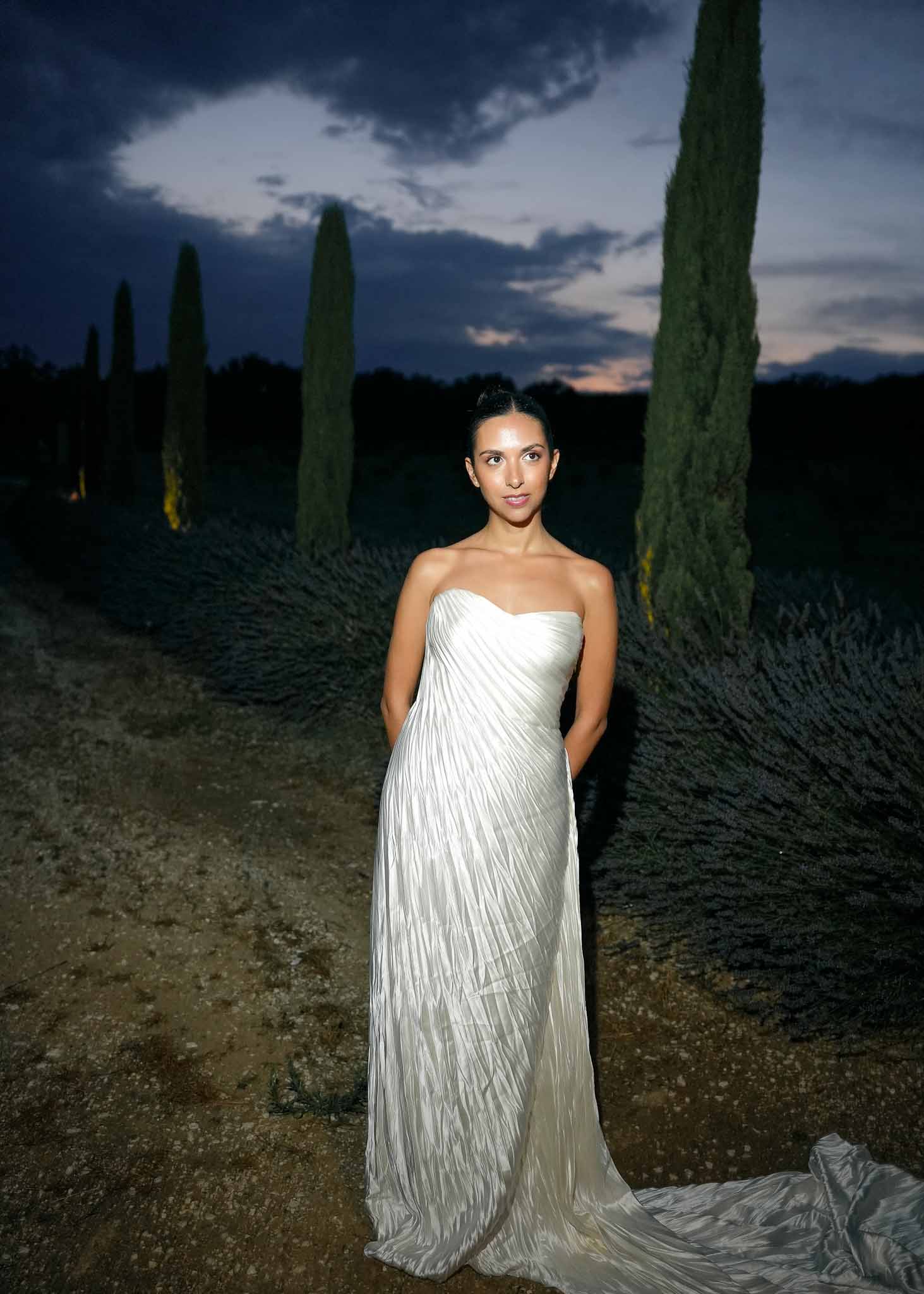 Bride in strapless white pleated column gown on dirt path with lavender and cypress trees at dusk