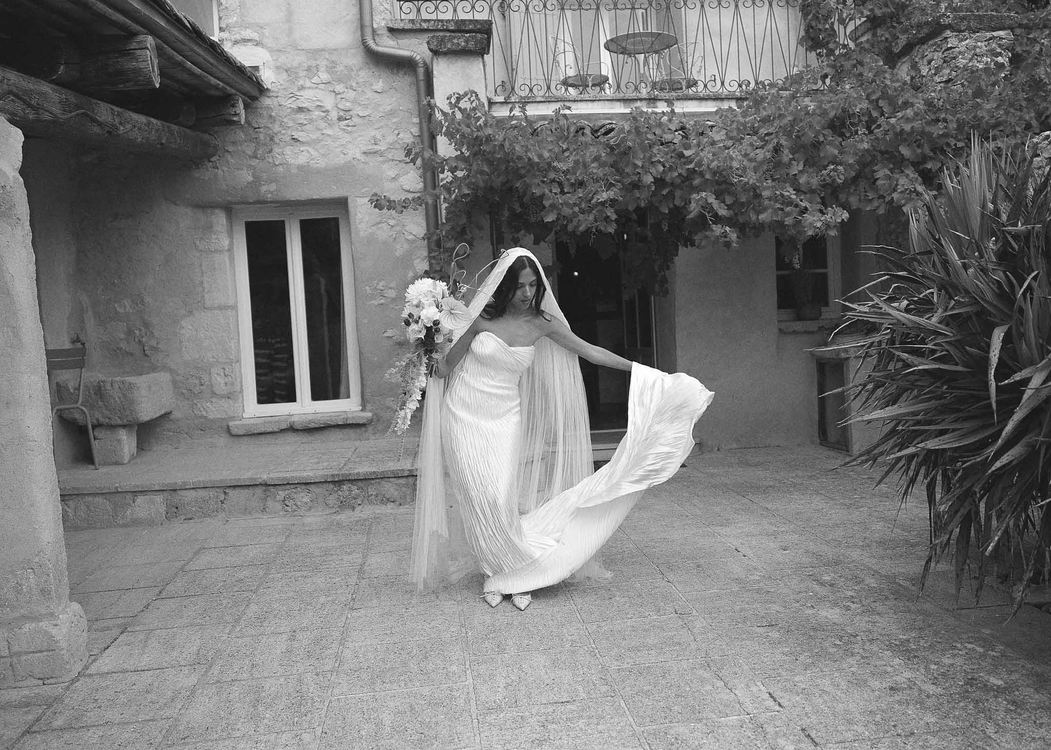 Black-and-white bridal portrait in stone courtyard with strapless pleated dress, billowing cathedral veil, and cascading b...