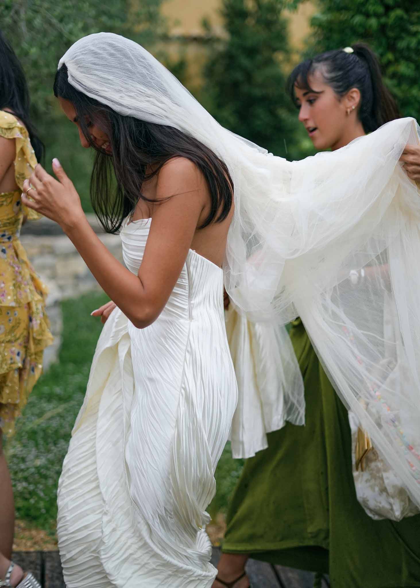 Bride laughing while bridesmaid arranges her veil walking through garden