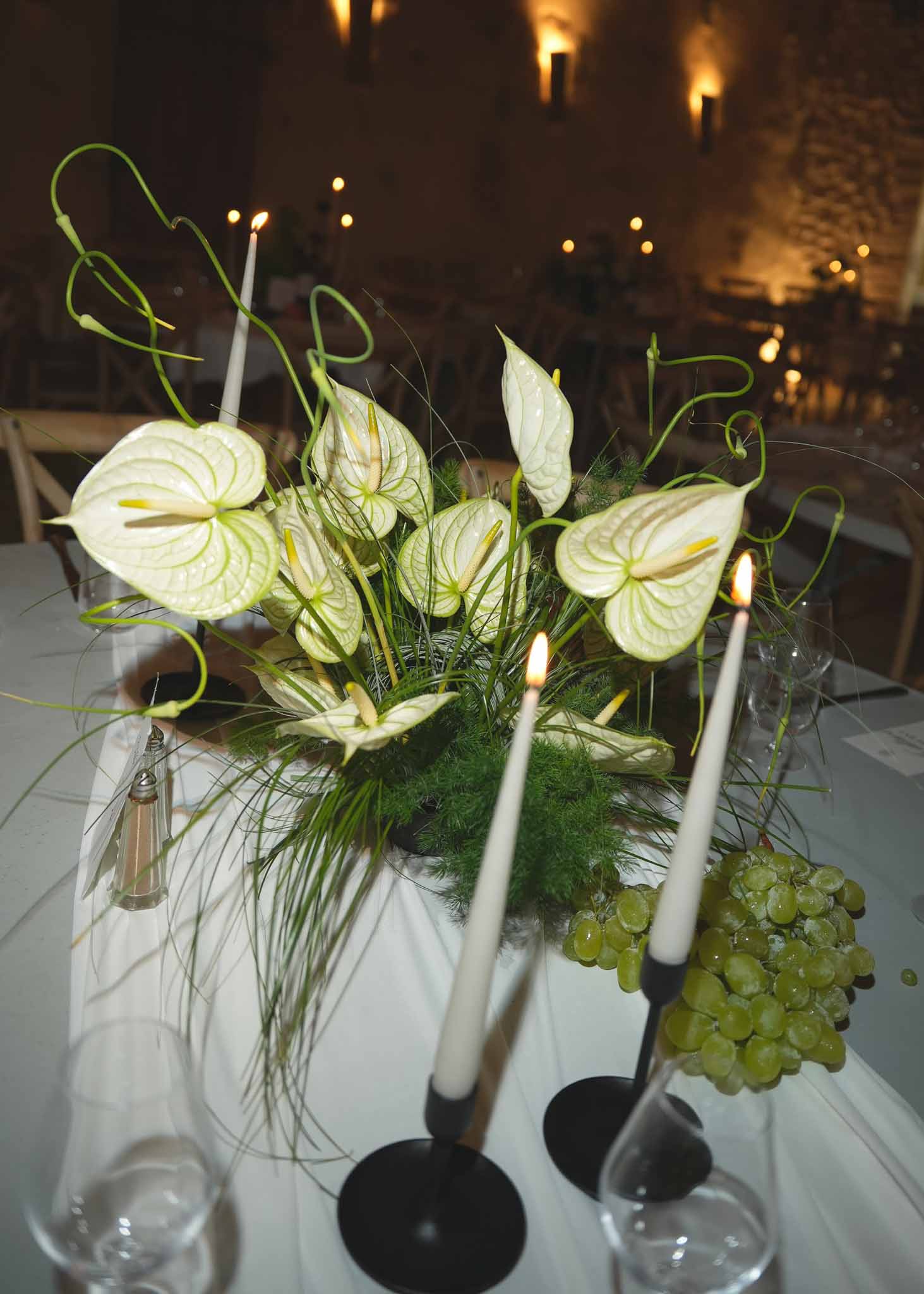 White-green anthurium centrepiece with fern and black taper candles beside grapes in stone-walled venue