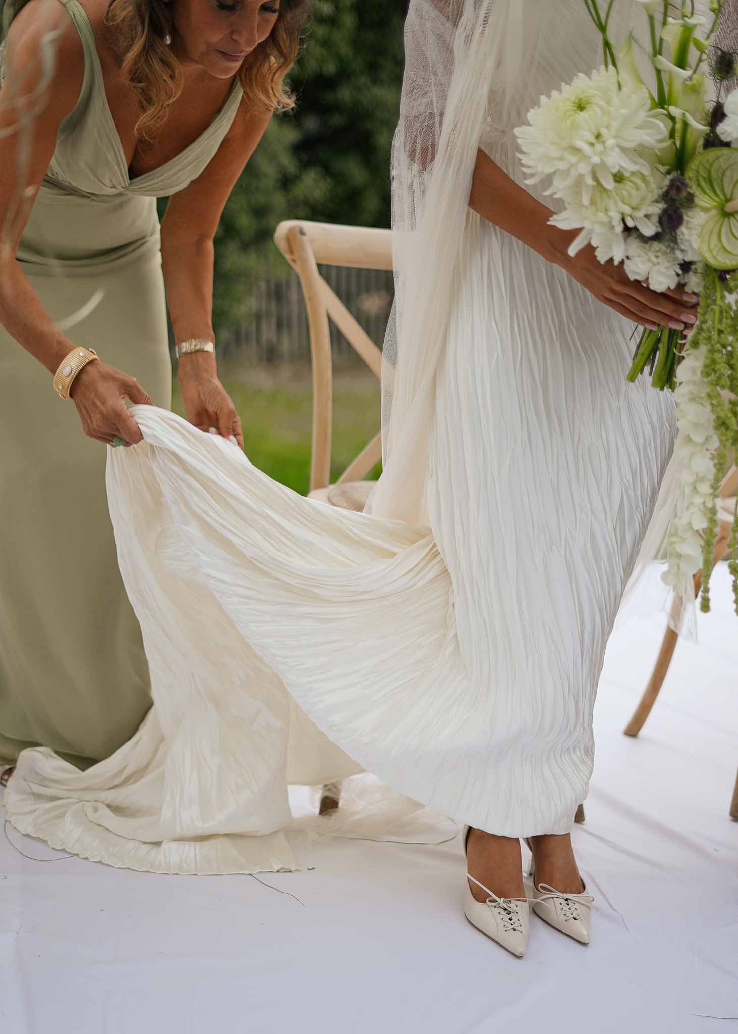 Bridesmaid in sage gown arranging bridal train with white dahlia and thistle bouquet visible