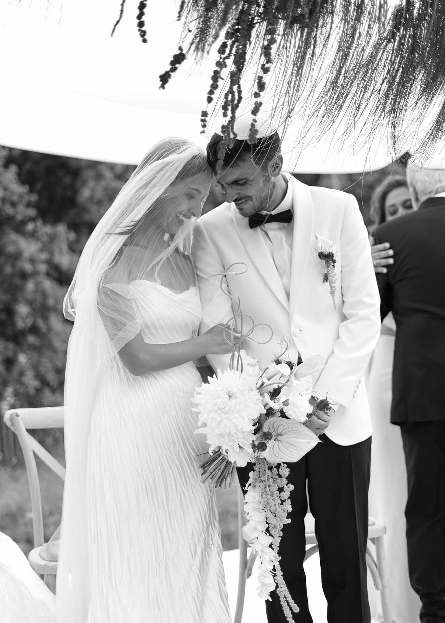 Black and white portrait of laughing bride and groom under dried palm frond canopy during outdoor ceremony