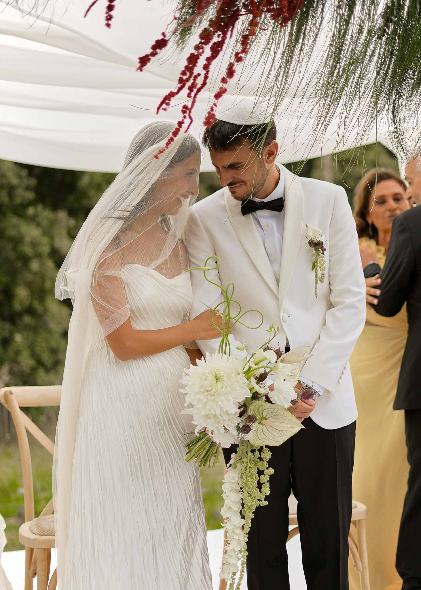 Couple under white chuppah with burgundy amaranthus, bride in pleated gown with cascading dahlia and hellebore bouquet