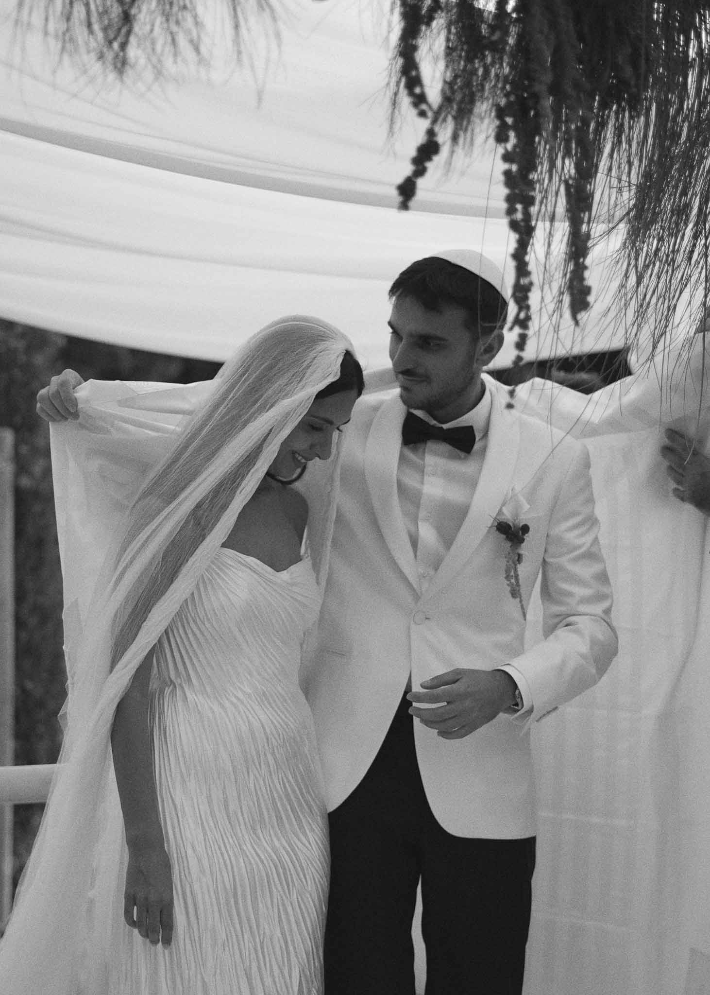 Black and white of couple under chuppah with trailing foliage, groom in kippah and white dinner jacket