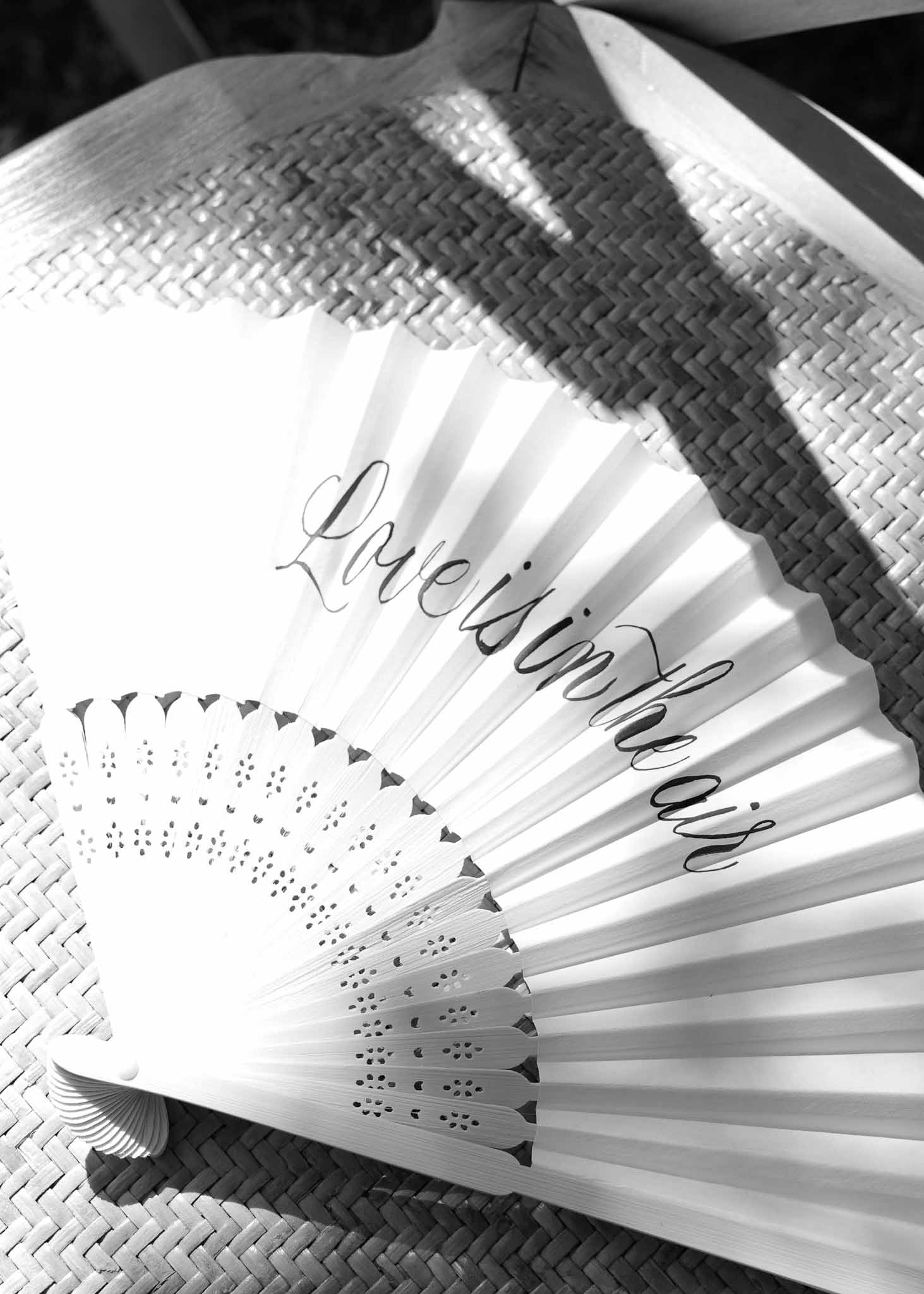 Black-and-white close-up of two white folding hand fans with calligraphy reading Love is in the air on woven straw surface