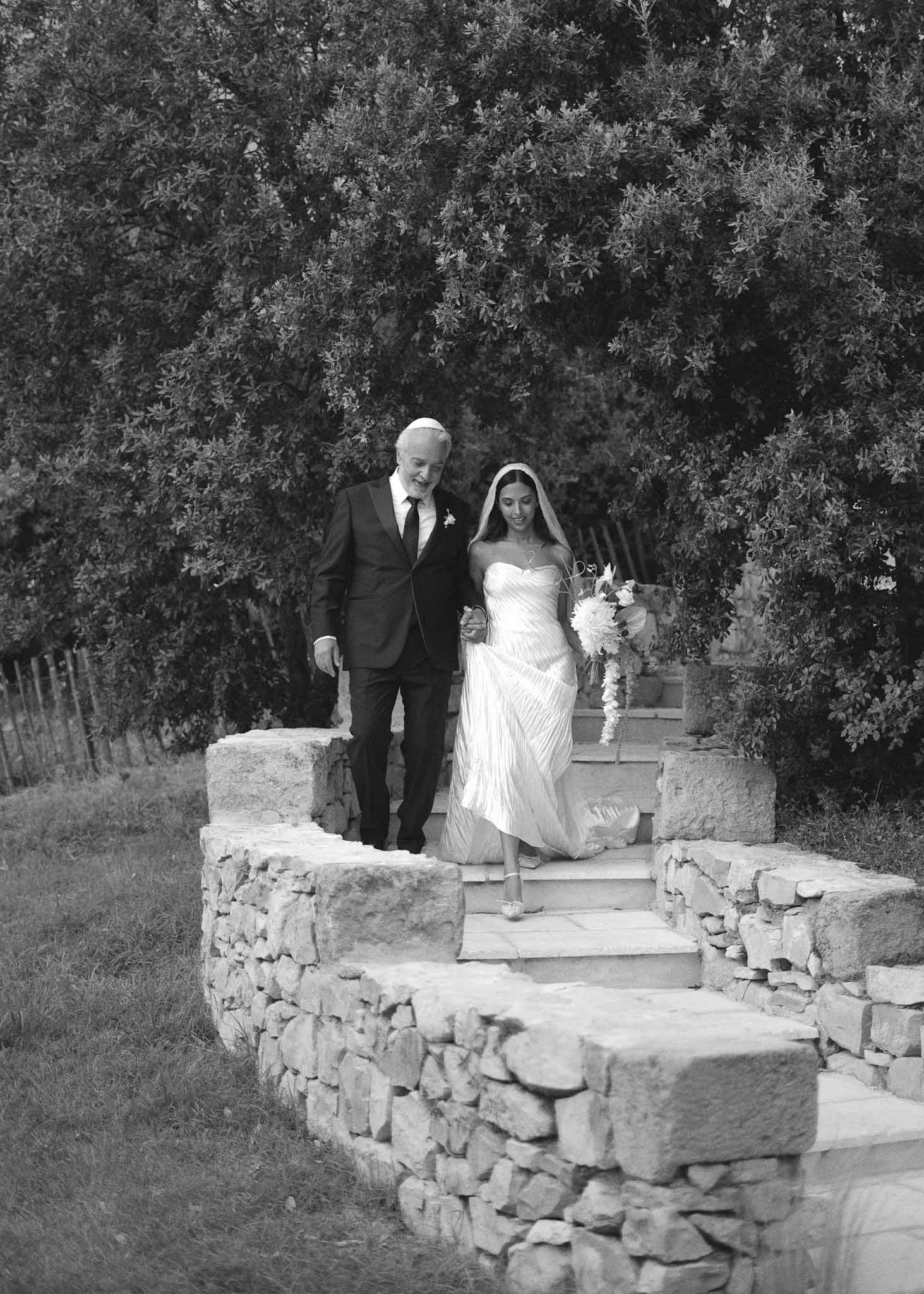 Black and white father walking bride down stone steps with vineyard in background