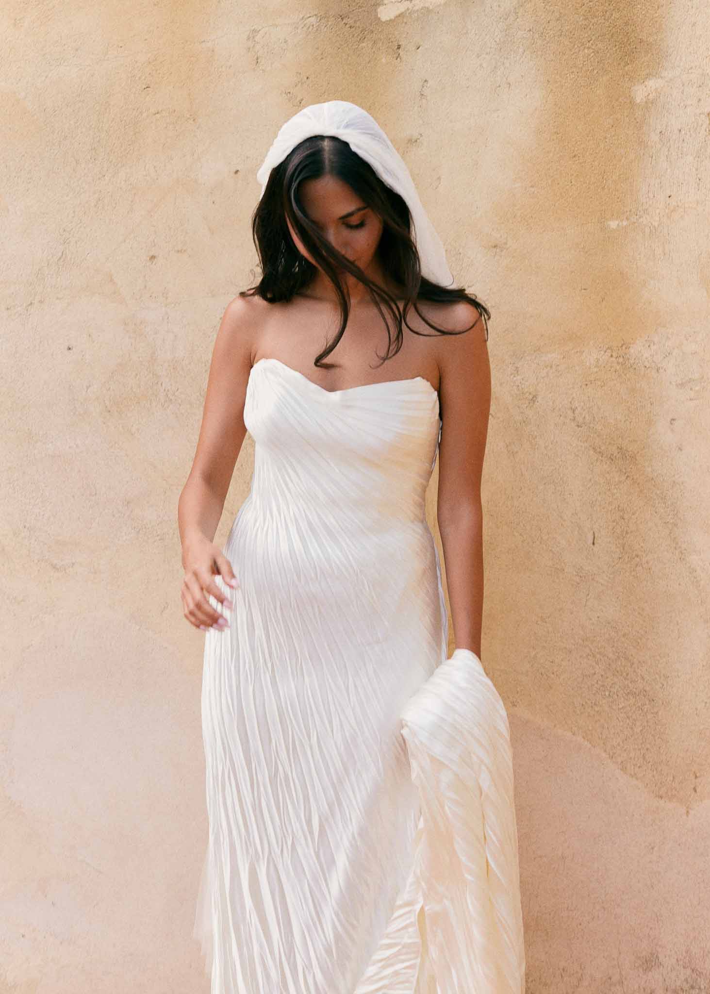 Bride in strapless white pleated gown with modern draped fabric headpiece against sandy-toned wall