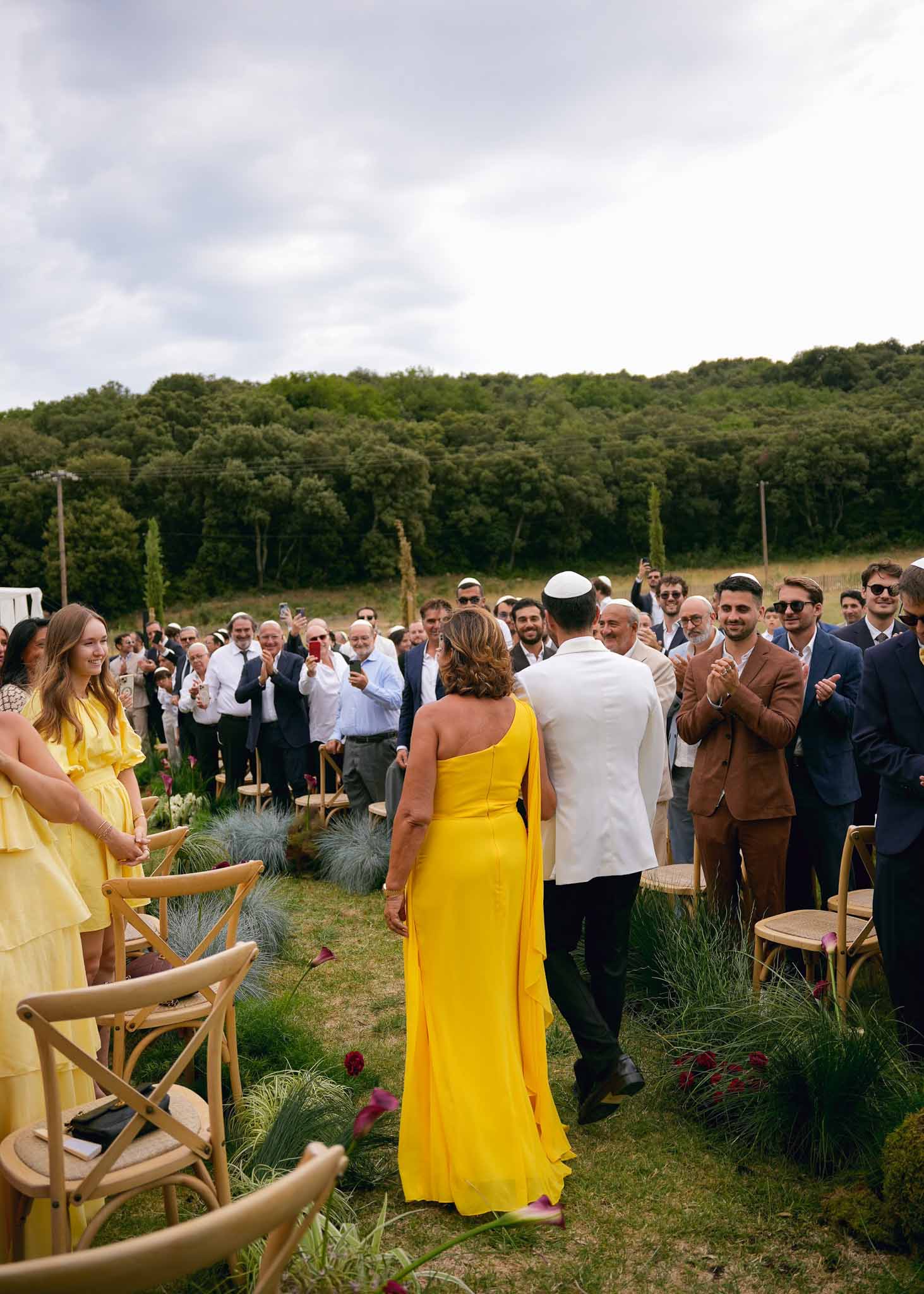 Groom in white jacket and kippah walking aisle with mother in yellow gown past burgundy floral arrangements