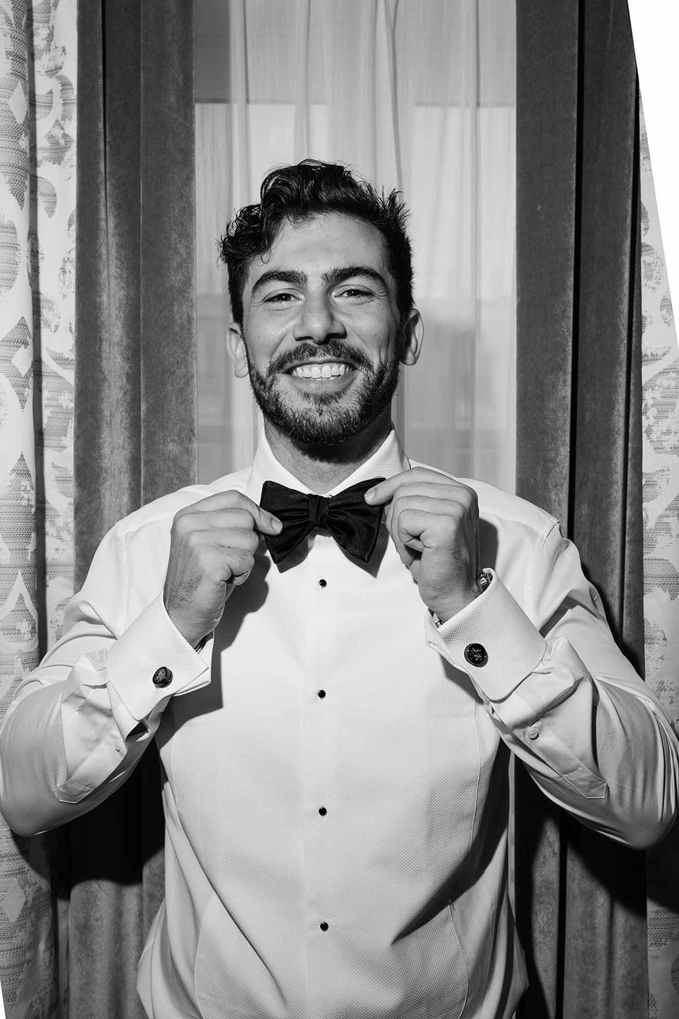 Black and white groom smiling and adjusting bow tie in white tuxedo shirt against patterned curtains