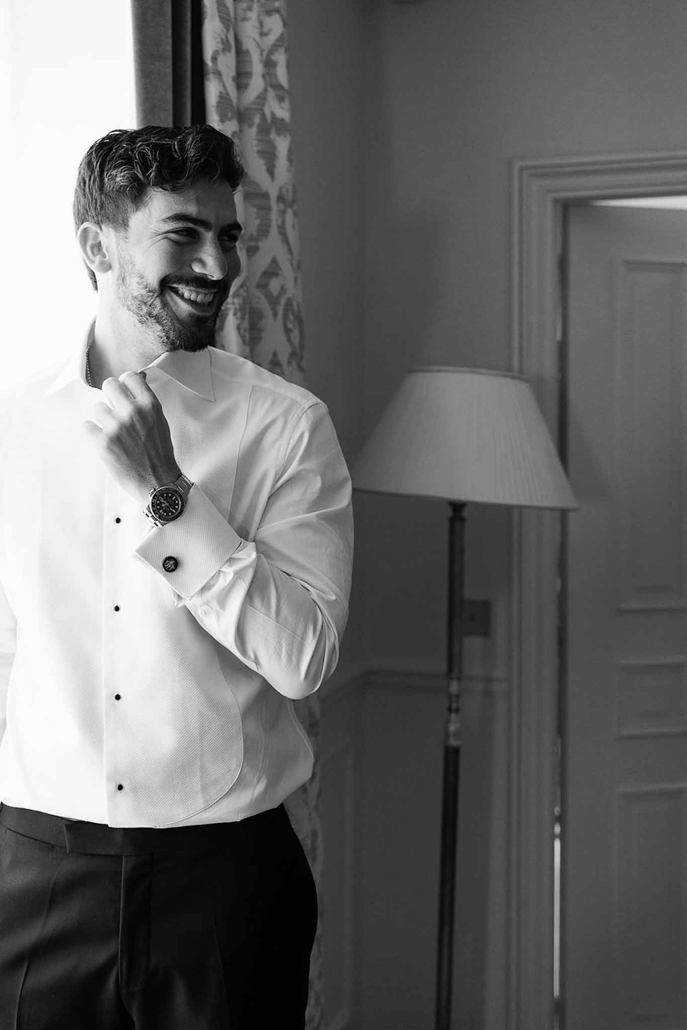Black-and-white portrait of groom laughing while adjusting dress shirt collar by window during getting ready