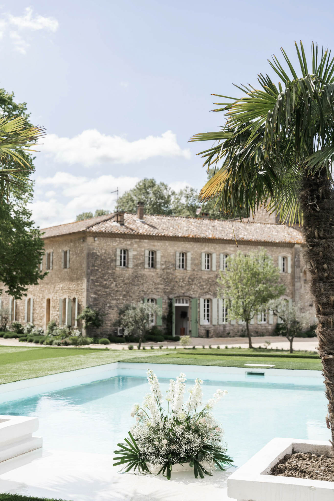 Provencal stone bastide with sage shutters behind swimming pool decorated with white orchid arrangement and palms