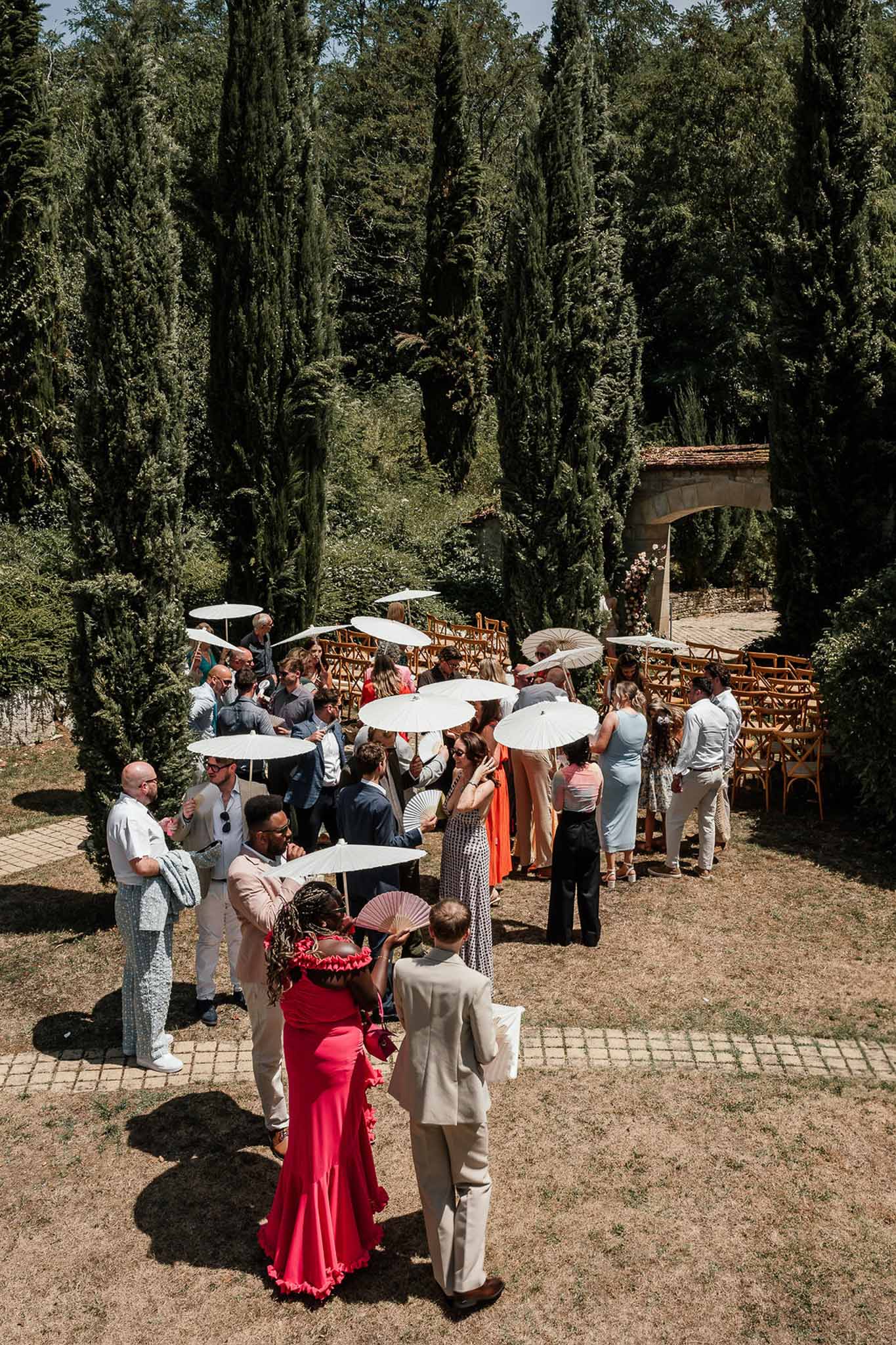 Guests with white parasols and fans gathering before outdoor ceremony with wooden chairs and floral arch
