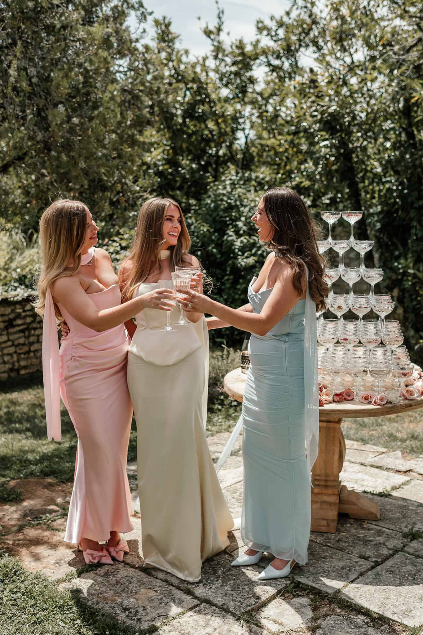 Bride and two bridesmaids in pink and blue satin dresses toast with coupe glasses beside champagne tower
