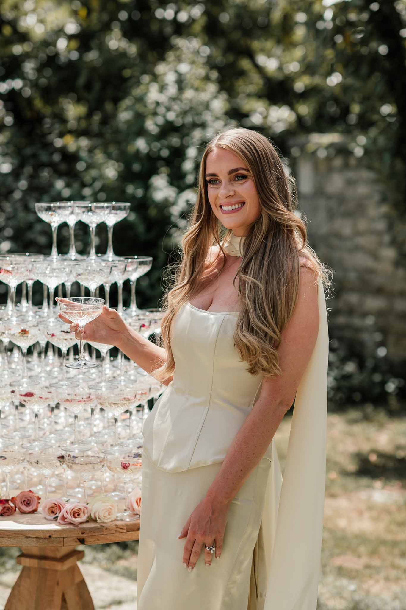 Bride in ivory strapless jumpsuit with cape raising coupe glass beside crystal champagne tower