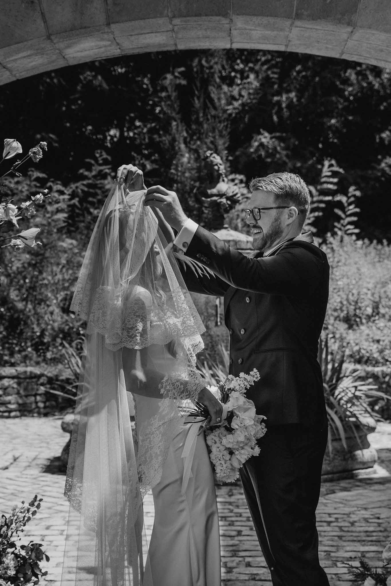 Black and white portrait of groom lifting lace cathedral veil over bride beneath stone arch