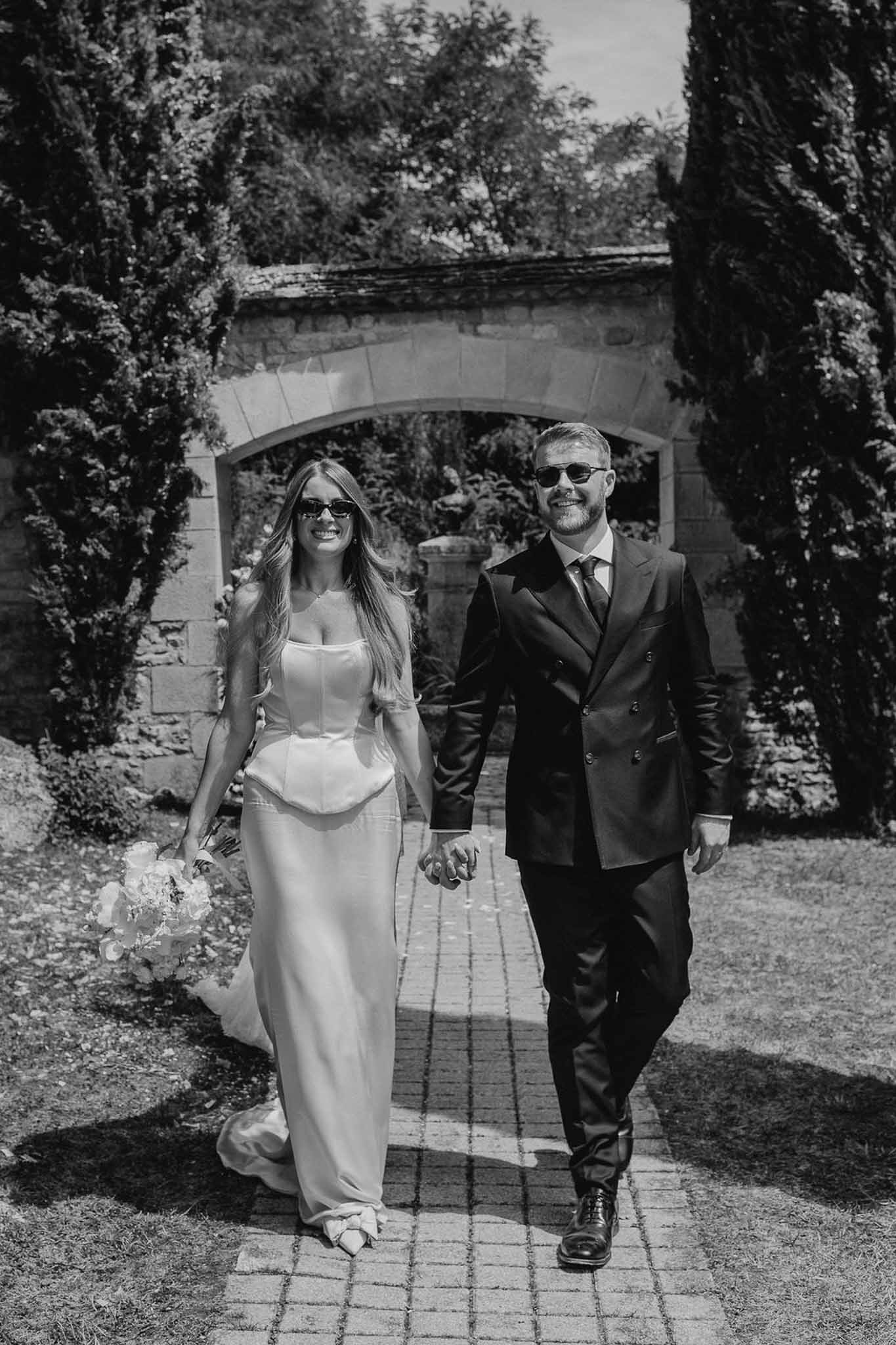 Black and white portrait of bride and groom in sunglasses walking hand in hand through stone archway with cypress trees