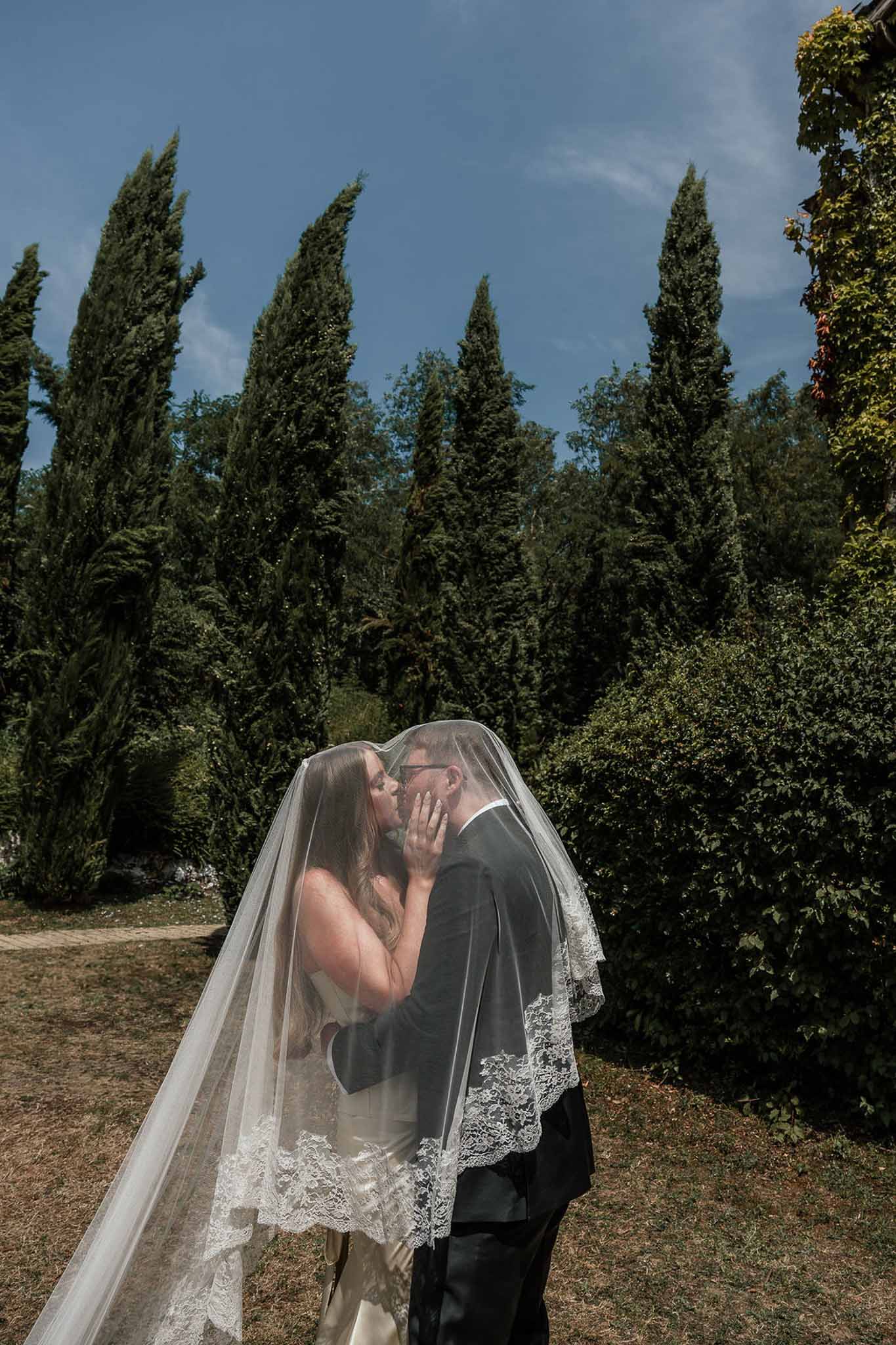 Couple kissing beneath lace-trimmed cathedral veil creating tent effect against cypress tree backdrop
