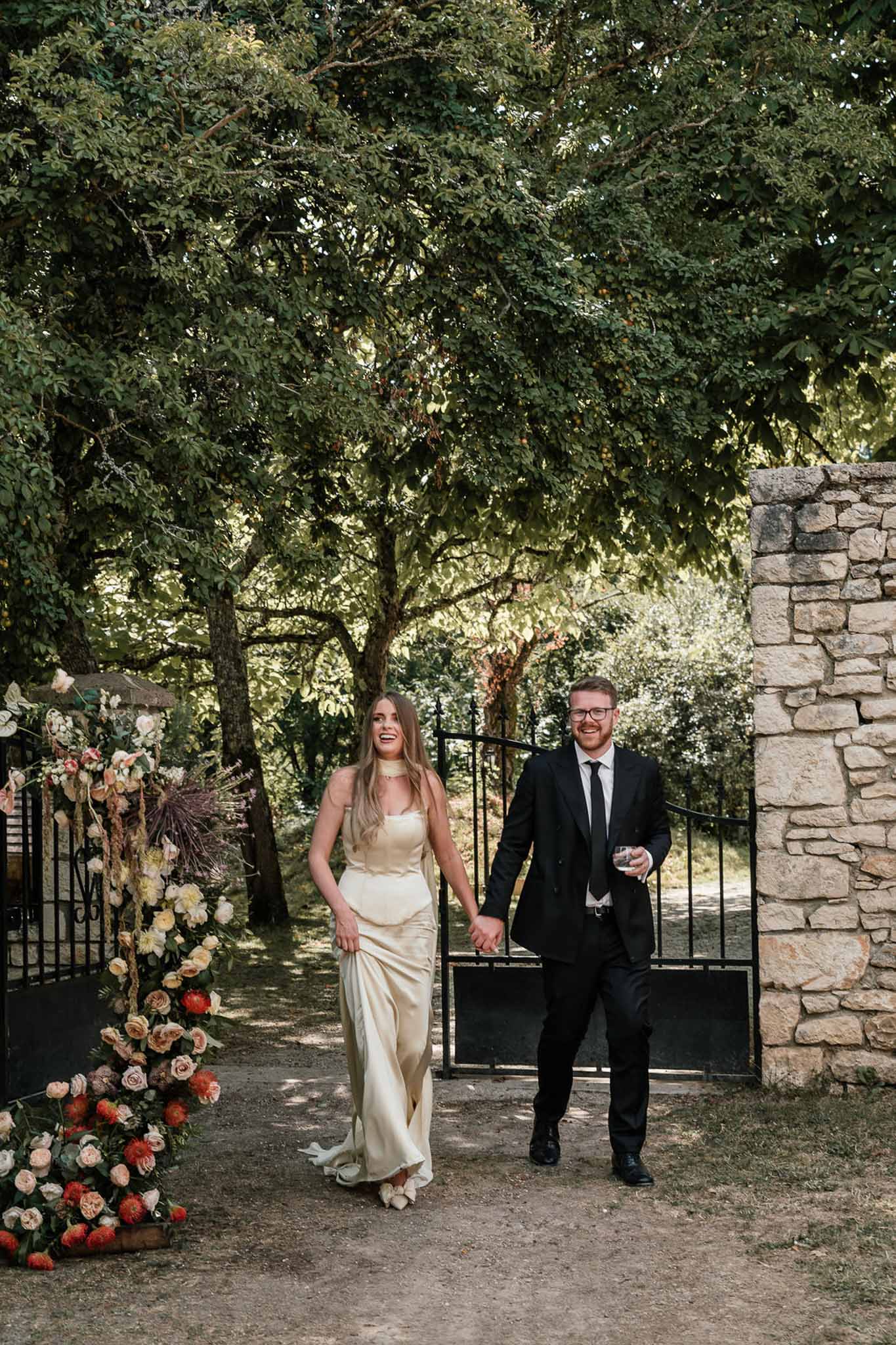 Bride in gold satin slip dress and groom in black suit walking through iron gate with coral floral arrangement