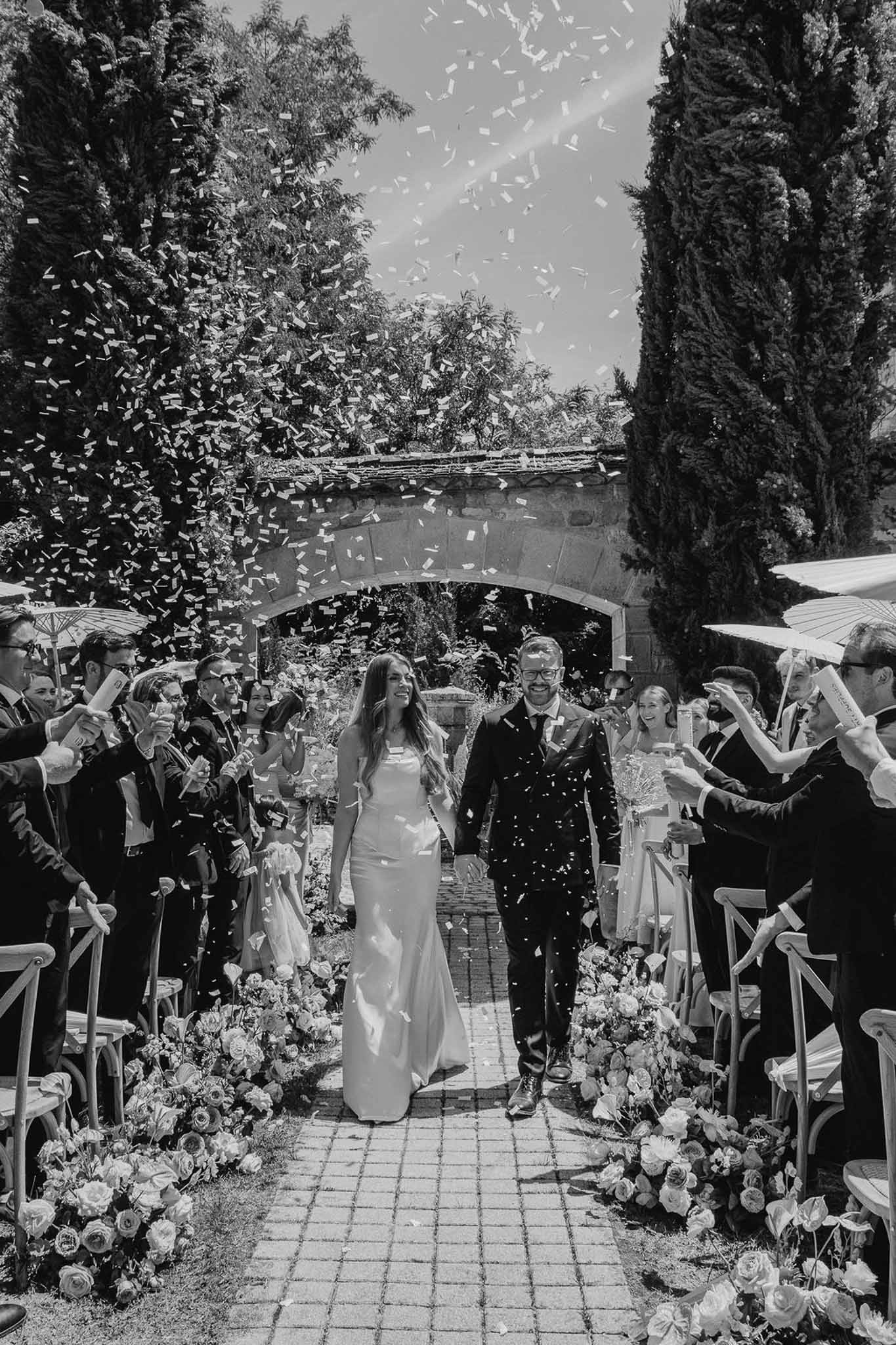 Black and white of couple exiting ceremony through confetti shower on brick aisle with stone archway and cypress trees
