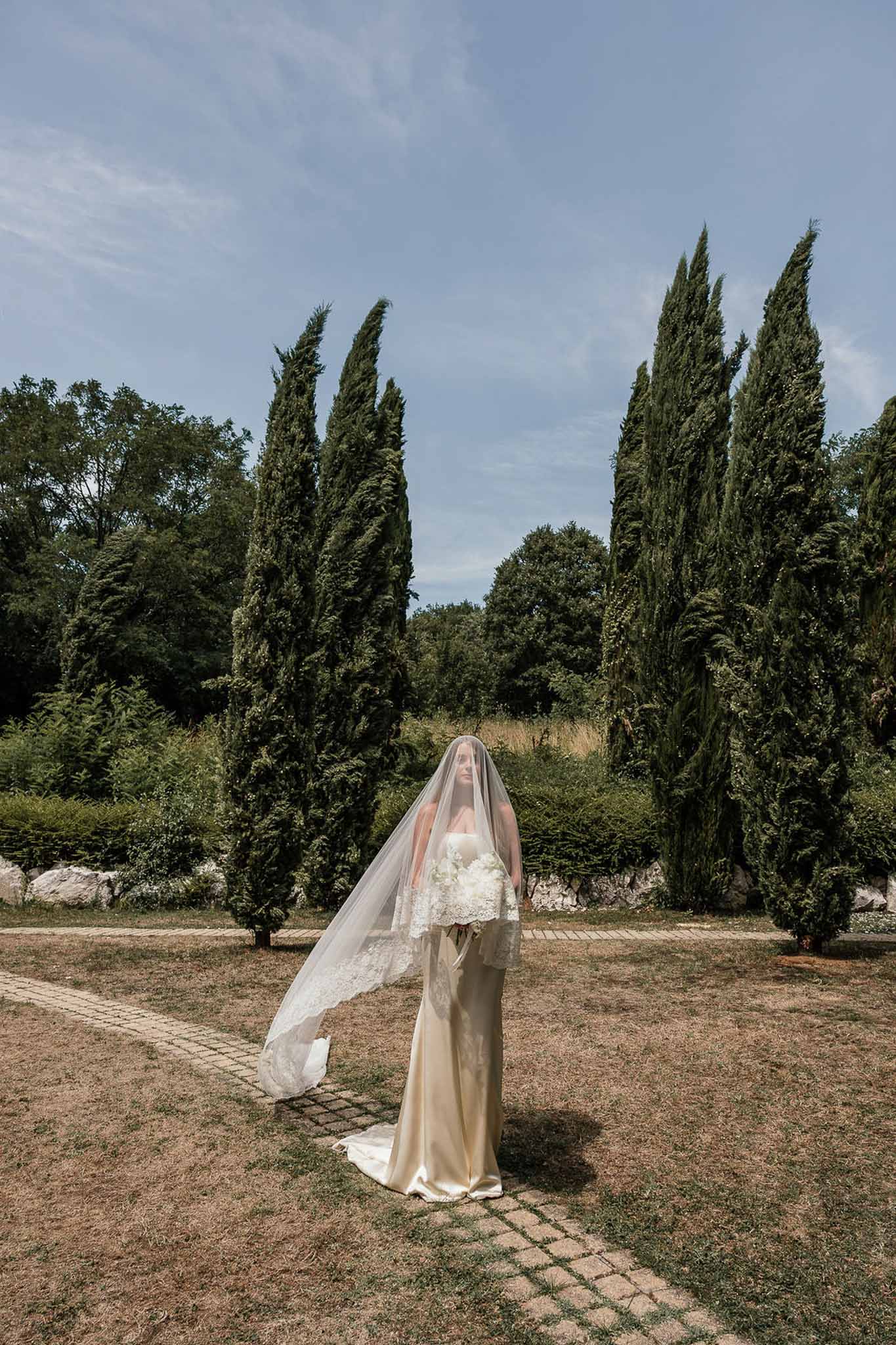 Bride in strapless satin gown with cathedral lace veil holding white bouquet on garden path with cypress trees