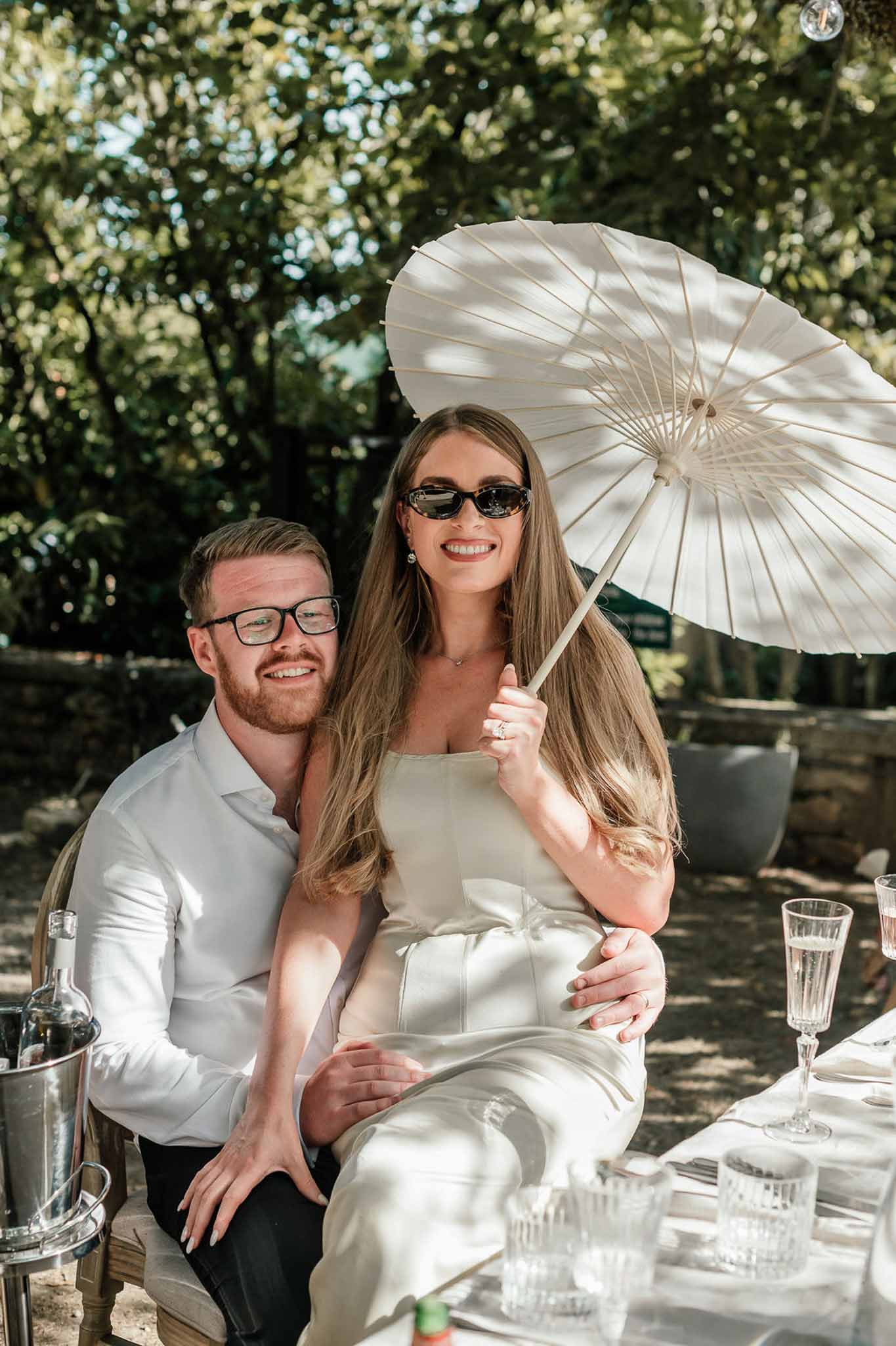 Bride in ivory slip dress with parasol seated on groom's lap at garden table with champagne glasses