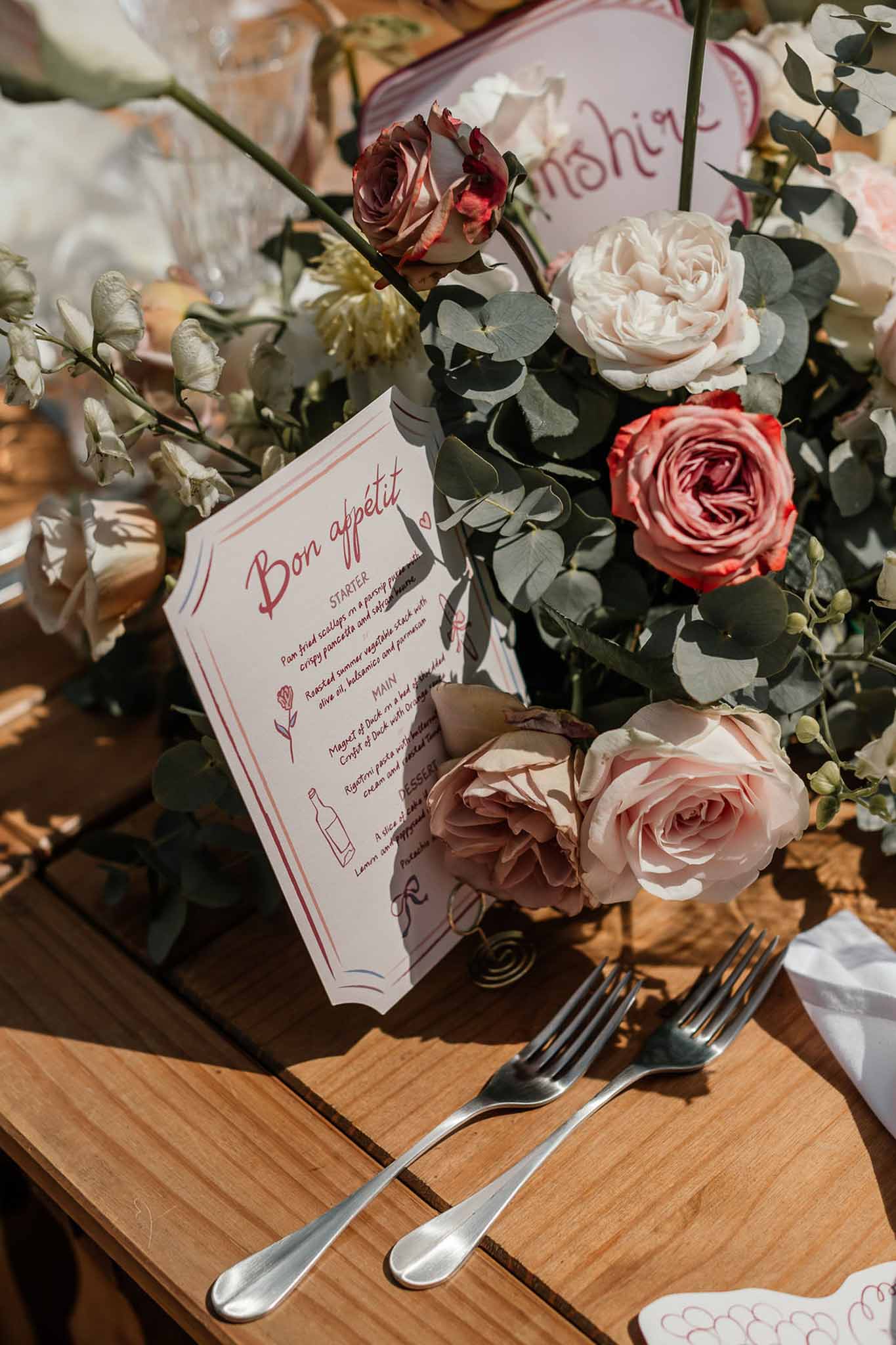 Wedding menu card reading Bon appetit surrounded by blush pink, burgundy, and cream roses on a wood table