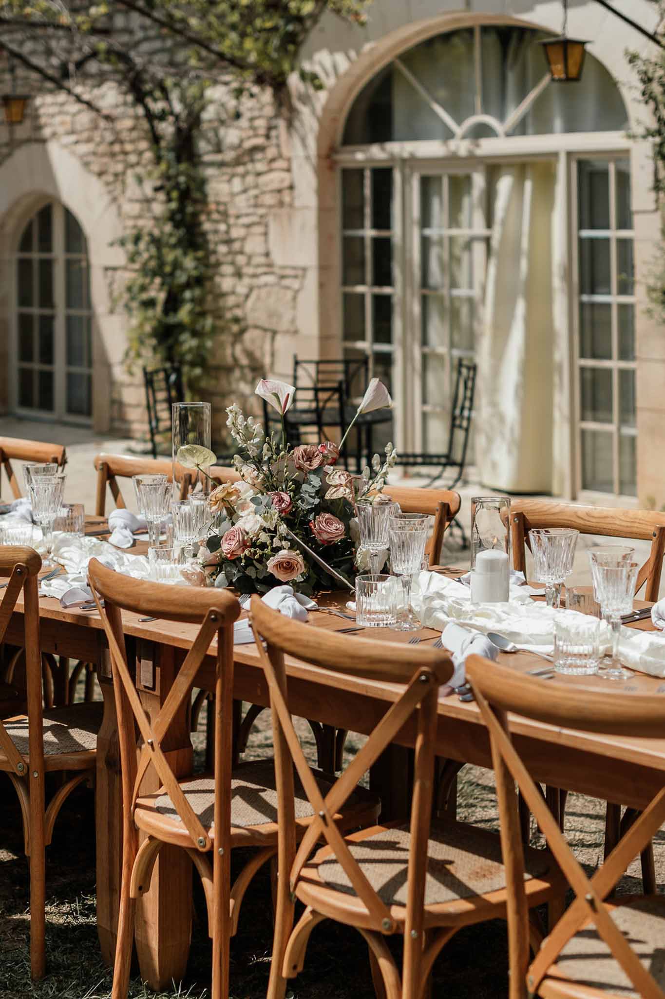 Long farm table set with cross-back chairs, pink roses, and candles in chateau courtyard with arched facade