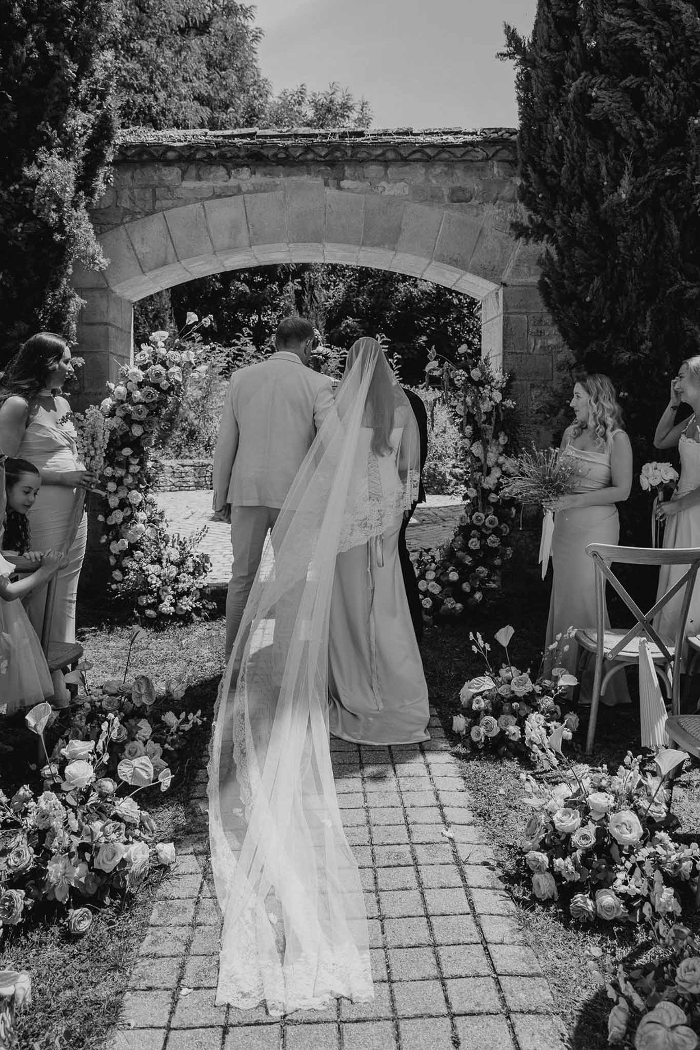 Black and white wide shot of bride and groom at stone arch ceremony with floral-lined aisle and cathedral veil