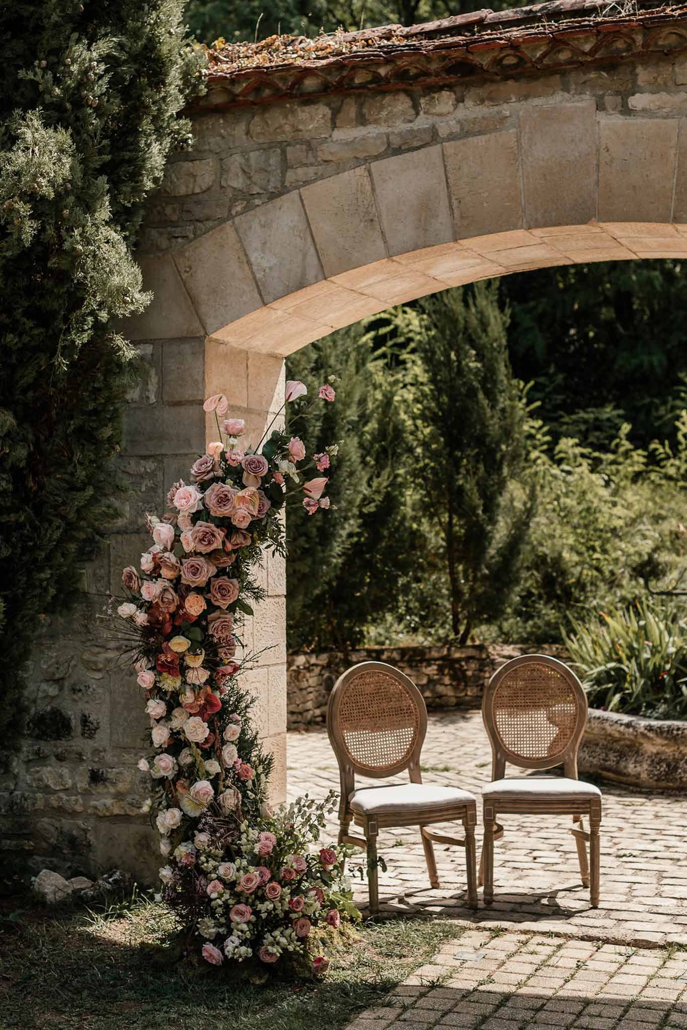 Outdoor ceremony chairs beneath stone archway with climbing floral installation of pink, mauve, and burgundy roses