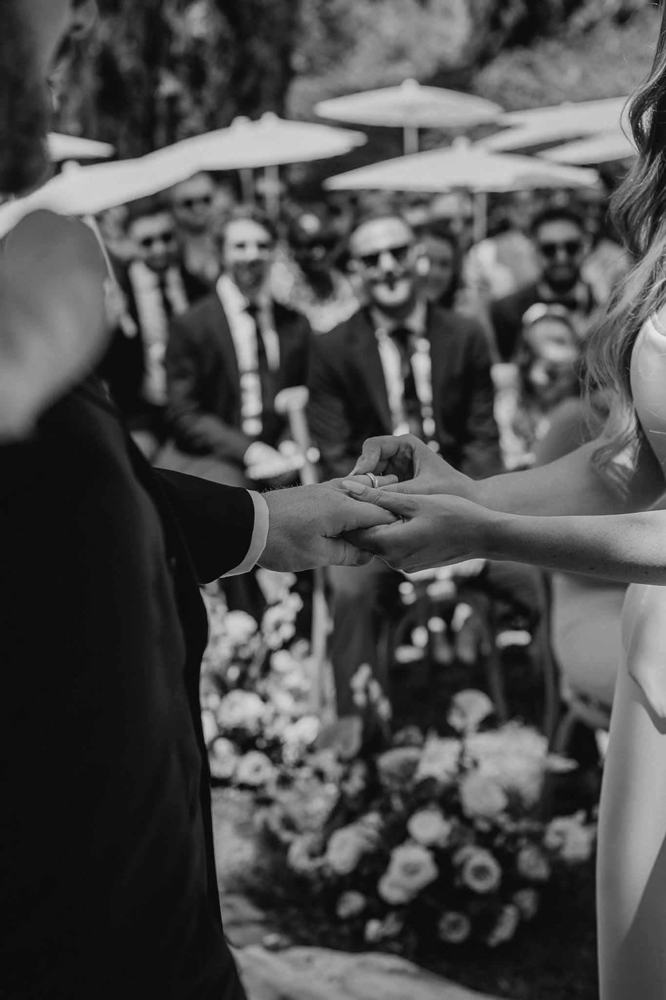 Black and white close-up of ring exchange with male guests in dark suits watching in background