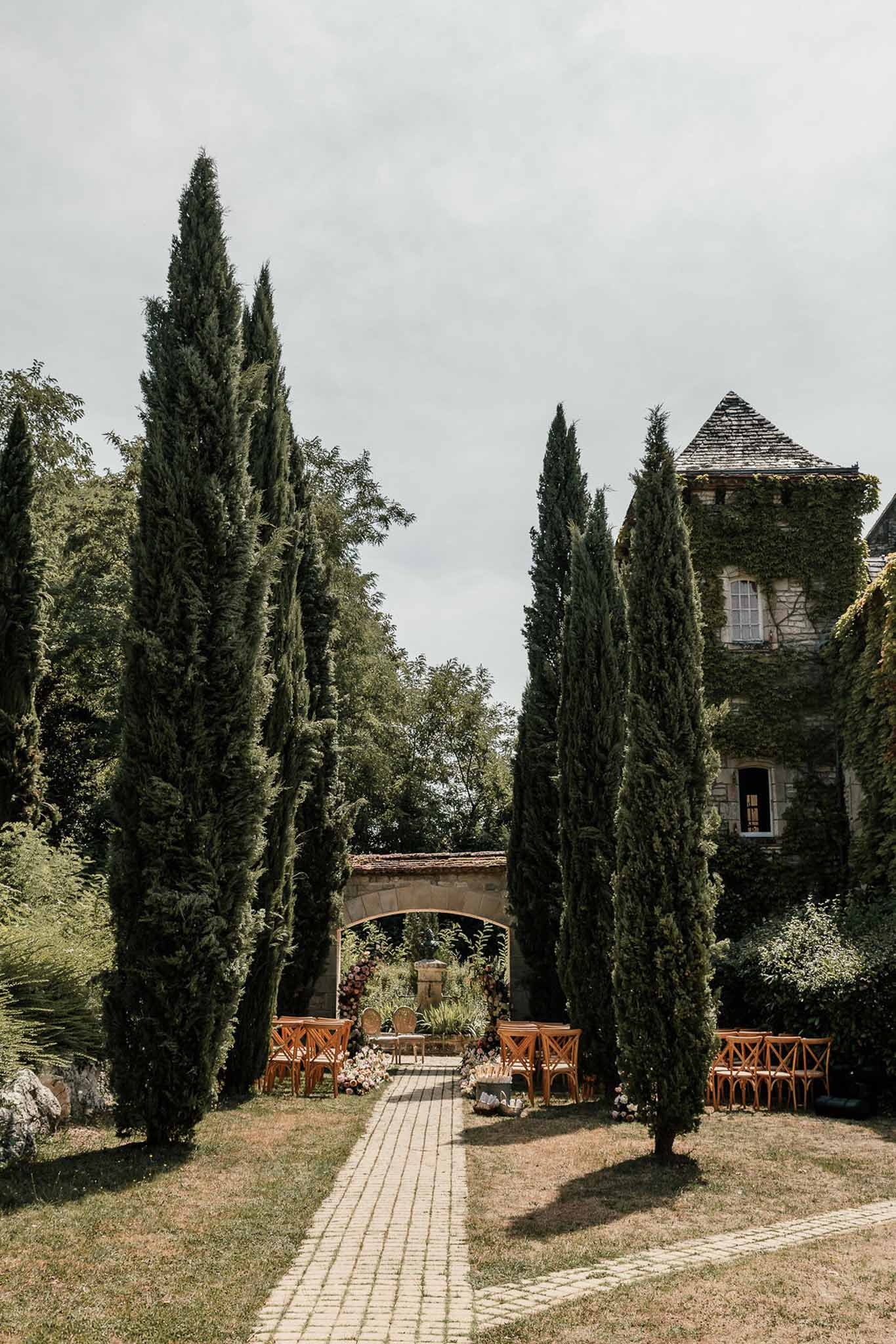 Ceremony setup with crossback chairs and floral arch flanked by cypress trees beside ivy-covered tower