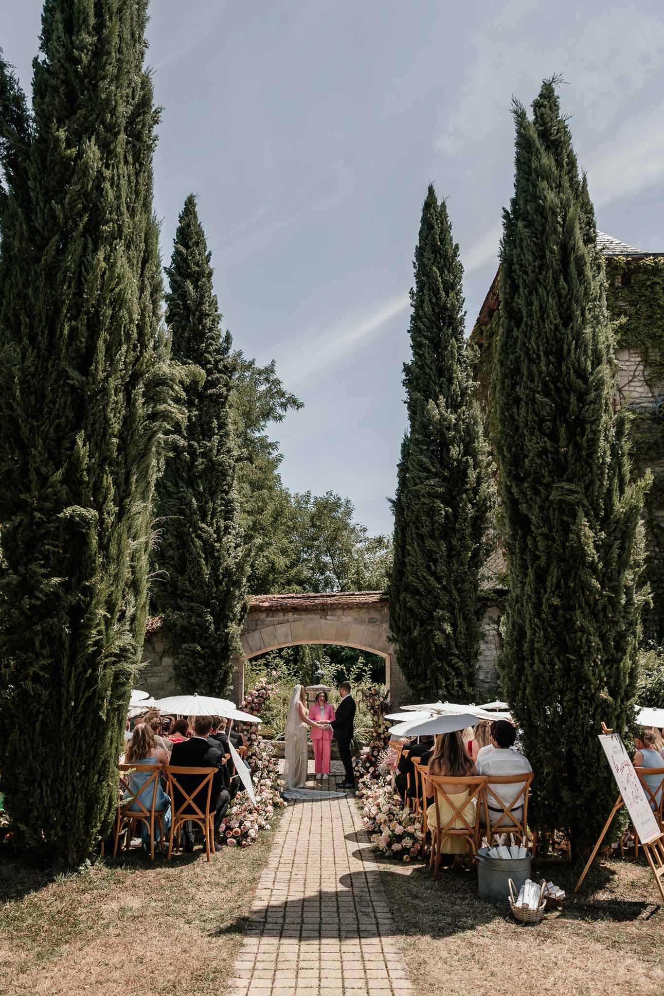 Ceremony in cypress courtyard with blush and cream floral clusters and 30 guests on crossback chairs