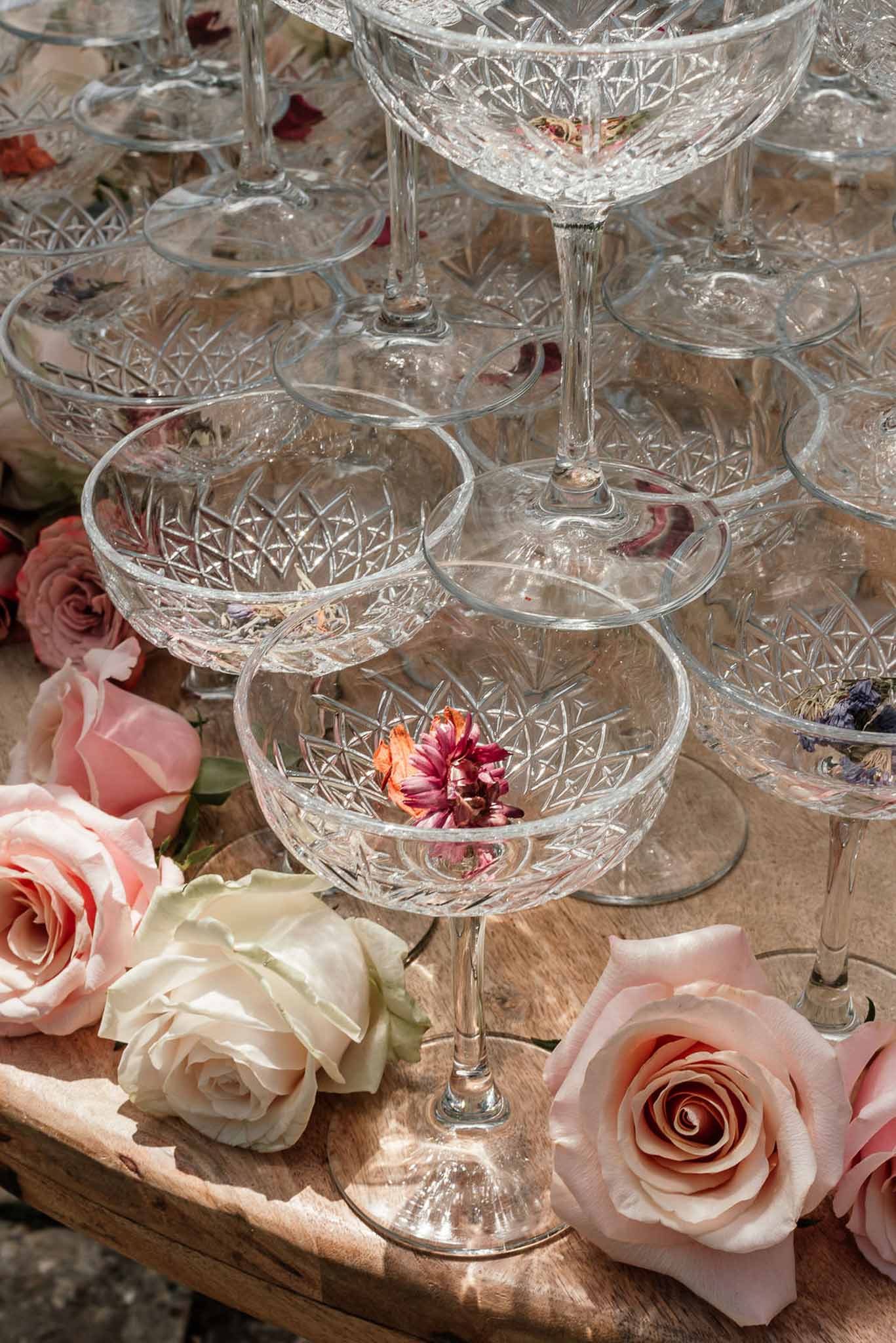 Crystal coupe champagne tower with dried burgundy blooms inside glasses and scattered rose heads on wooden surface