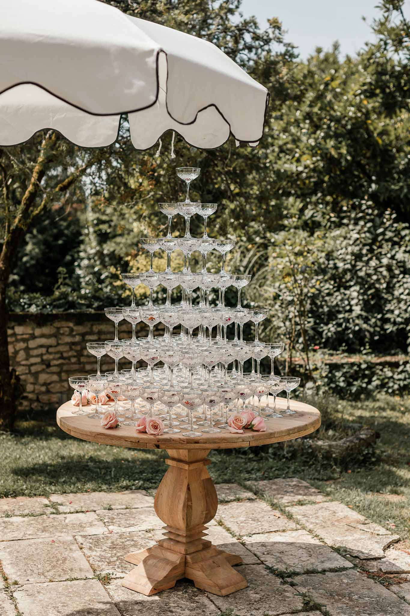 Seven-tier champagne coupe tower on wood pedestal table with scattered blush roses on stone terrace